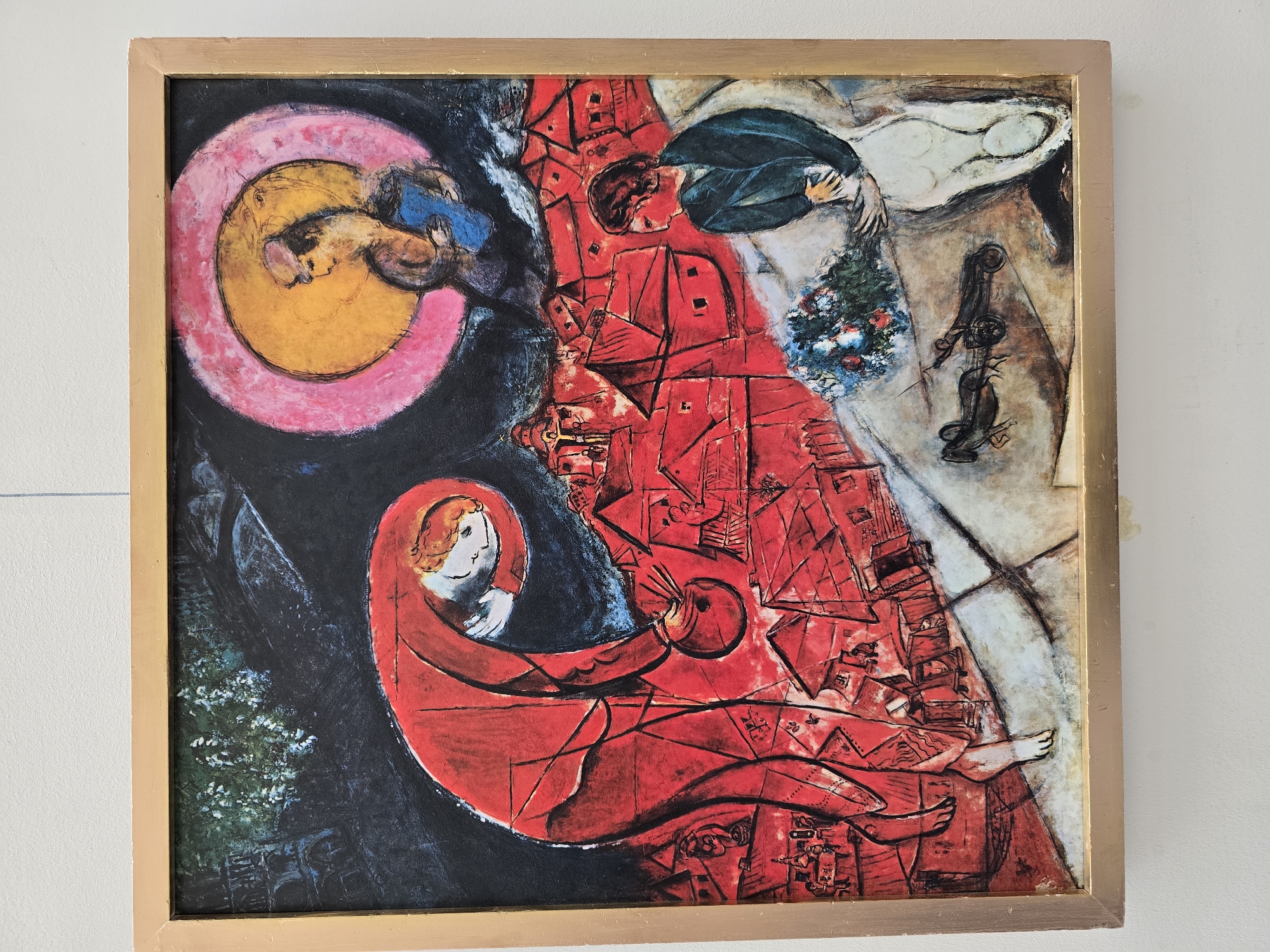 This is a painting by Marc Chagall depicting a dreamlike scene with figures and a cityscape. The dominant colors are red, yellow, and black. In the foreground on the left, a large, elongated figure in a red garment bends over, holding a circular object, possibly a lute, near a cluster of red-roofed buildings. The buildings appear to be stylized, with some featuring tiny figures. In the upper right, a circular halo or sun, colored yellow and pink, encloses two figures embracing. The figure on the right of the halo is holding a blue object. Below and to the right of the main cityscape, a figure in a dark jacket and white trousers looks towards the left. In front of this figure is a bouquet of flowers and a small cart pulled by a horse. The background includes dark trees and what appears to be a bridge or architectural structure on the far left. The painting evokes a sense of surrealism and emotional intensity, characteristic of Chagall's work. The artist was born in the town of Vitebsk, Belarus, near Minsk, and his childhood experiences in Belarusian Jewish shtetls often inspired his artwork.