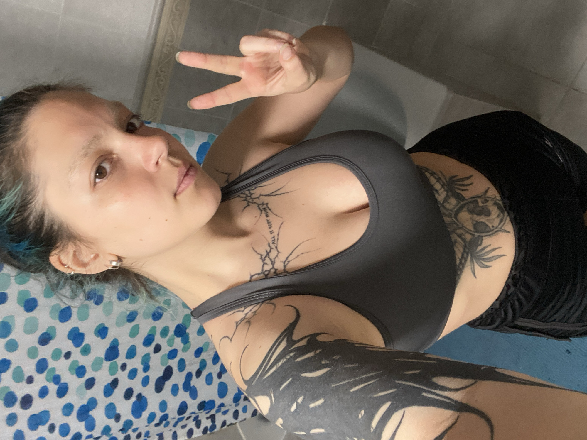 The rotated image captures a young adult female posing for a selfie indoors. She has brown eyes, light skin, and dark hair with distinct blue highlights visible near her left ear, which is adorned with multiple hoop piercings. She is dressed in a grey sports bra and dark, possibly black, athletic shorts. Her right hand is raised, making a peace sign (V-sign) towards the camera.
She features several prominent dark tattoos. A detailed abstract design resembling roots or branches spans her upper chest, with the text "ALL IT IS OFT MORE" legible within it. Her right arm and shoulder are extensively covered by a large, dark, tribal-style or abstract tattoo. On her left side, partially visible below her sports bra, is another dark tattoo depicting palm trees and other elements.
The background shows a light-colored tiled wall with a decorative upper border. To her left, a patterned surface with blue and light blue dots is visible, possibly a sheet or mat. To her right, a plain blue surface is present, likely a mat. Her expression is neutral and direct, suggesting she is aware of the camera. The setting is indoors, in Gijón/xixón, Spain.