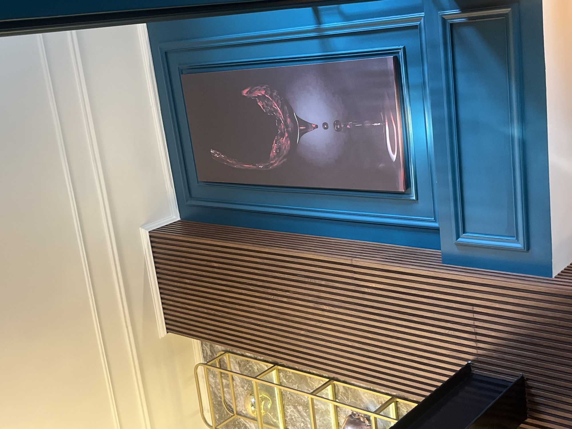 The image shows a section of a room with a modern, stylized design. There is a blue-framed art piece depicting a wine glass with liquid in motion. Below the art, a wall decorated with horizontal wooden slats is visible. A gold-colored metal rack with decor items is also present. Part of a white wall with molding is seen on the left. Based on the visual style and color palette, the image could be from an upscale restaurant or bar in a cosmopolitan area; however, the provided context of Puebla, Mexico, suggests this could be part of a modern establishment blending local elements with contemporary design.