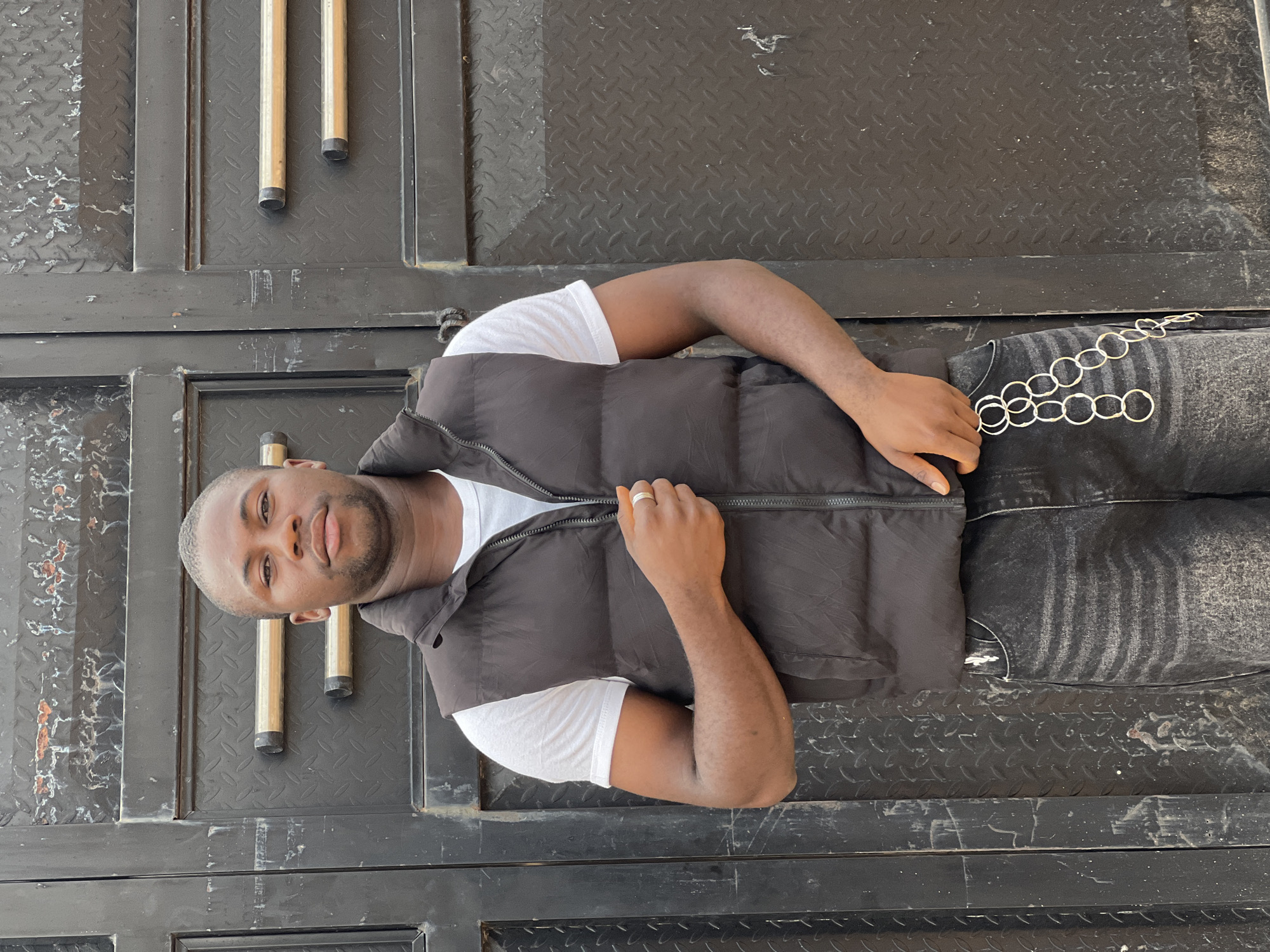 A light-skinned man is laying on his side on a black metal surface in Bukuru, Nigeria. He is wearing a white t-shirt, a black vest, and dark jeans with a silver chain hanging from his waist. He is looking at the camera. The background is a black metal surface with a diamond plate pattern. To his left is a black metal door with two vertical gold handles. There is some light-colored residue on the metal surface.