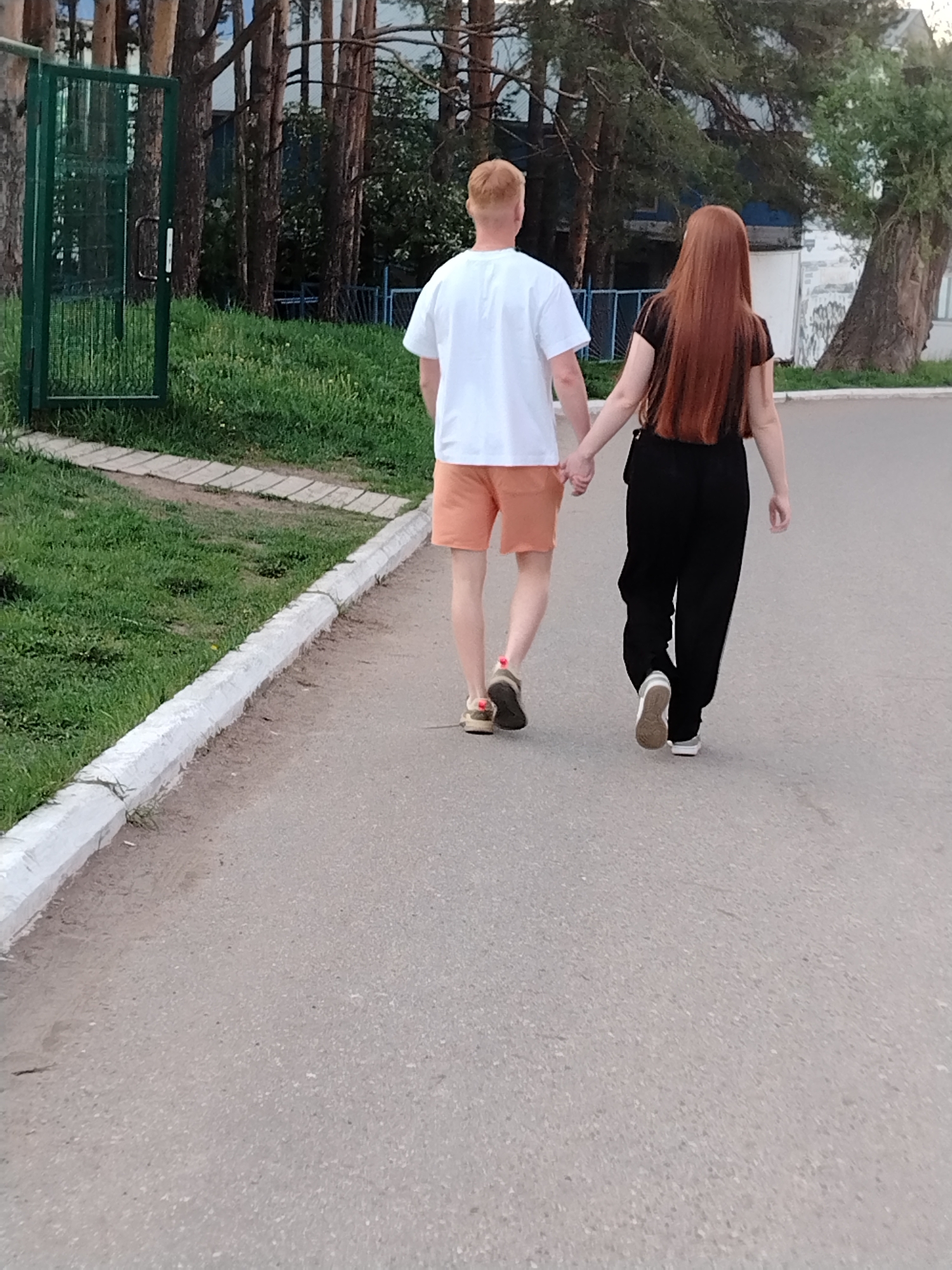 A couple is walking hand-in-hand on a paved road in Chaikovsky, Russia. The man has short red hair, a white t-shirt, and orange shorts. The woman has long red hair, a black shirt, and black pants. They are walking away from the camera. A green metal gate and several pine trees are visible in the background. A grassy area with a white curb is to the left of the road.