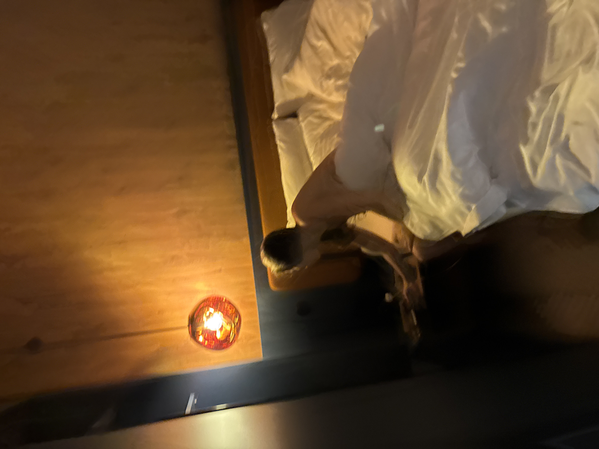 The image presents an overhead, angled view of a dimly lit bedroom scene, likely at night. The primary subject is a person sleeping or resting in a bed, largely covered by a rumpled white duvet or blanket. Their bare arm, shoulder, and upper back are visible, suggesting they are lying on their stomach or side, with their head turned away from the camera. The person has short, dark hair.
The bed is furnished with white pillows and matching bedding. Adjacent to the bed, a warm-toned wooden surface, possibly a bedside table or part of the floor, is illuminated by a decorative light source. This light source is a spherical, red or orange glass fixture with a visible, glowing filament bulb, casting a warm, soft light across the wooden surface. Below this wooden surface, a dark, featureless area is visible, which could be part of the bed frame, a lower shelf, or the floor.
The lighting throughout the scene is artificial and warm, contributing to a cozy and intimate ambiance, typical of a bedroom environment during evening or nighttime hours. There is no visible text in the image. The slightly blurry quality suggests low light conditions or slight camera movement during capture.