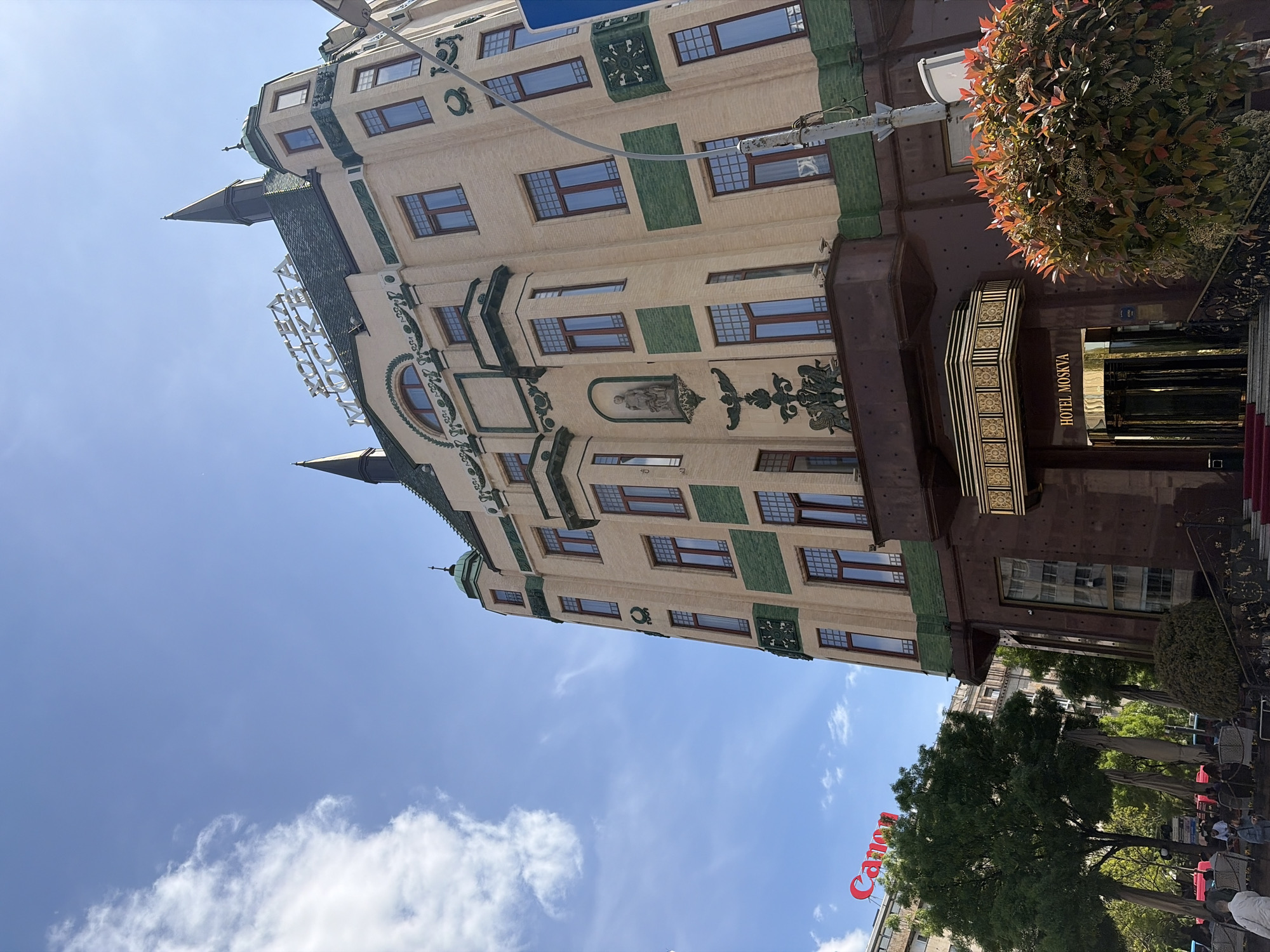 The image depicts the Hotel Moskva, a historic building in Belgrade, Serbia, angled upwards from a street-level perspective. The hotel is a multi-story structure with a distinctive facade, featuring ornate architectural details, numerous windows with brown frames, and green decorative elements. The hotel's entrance is visible, with a dark brown overhang and the name "HOTEL MOSKVA" displayed vertically. A red carpet is partially visible near the entrance.
In the foreground, parts of trees are present on the right and at the bottom. In the lower left is a portion of a building with the Canon brand partially visible. The sky is predominantly blue with scattered white clouds. The photo was taken during daytime.