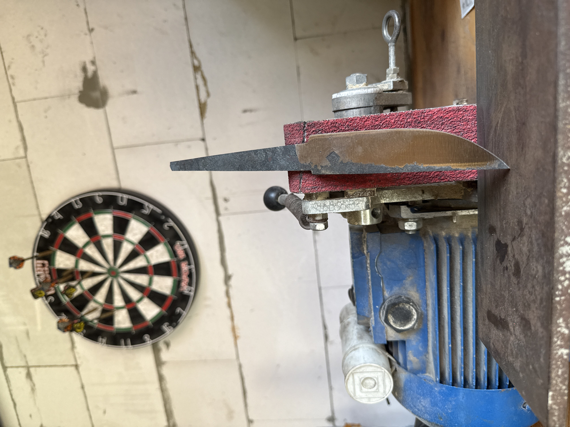 The image shows a knife being sharpened on a belt grinder in a workshop. A dartboard with several darts is mounted on a white brick wall in the background. The workshop appears to be in Krutaya, Russia, as indicated by the presence of the specific brand of darts. The lighting suggests an indoor setting, possibly a garage or a dedicated workspace. The overall atmosphere is one of practical application and potentially a hobbyist's setup.