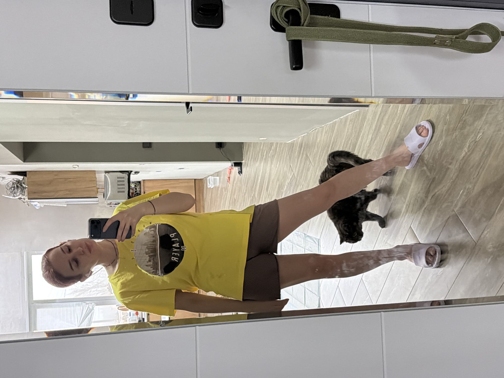 The image shows a young woman taking a selfie in a mirror. She is wearing a yellow t-shirt with a graphic print, brown shorts, and white slides. Her legs appear to have white paint on them. There is a black cat near her legs. The background appears to be an apartment interior, potentially a hallway or a living space with light-colored tiled floors. In the reflection, a partial view of a kitchen area with wooden cabinets and other household objects is visible. A green strap hangs on a white door or cabinet. The location is purportedly in the city of Chaikovsky, Russia.