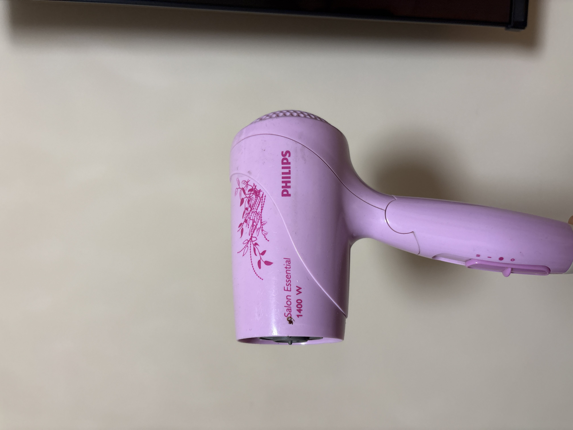A close-up shot depicts a light purple Philips Salon Essential 1400 W hair dryer. The dryer is positioned in front of a beige wall, with a dark object visible in the background at the top. There are no people in the image. The location in the city of Sofia, Bulgaria is not determinable from the image.