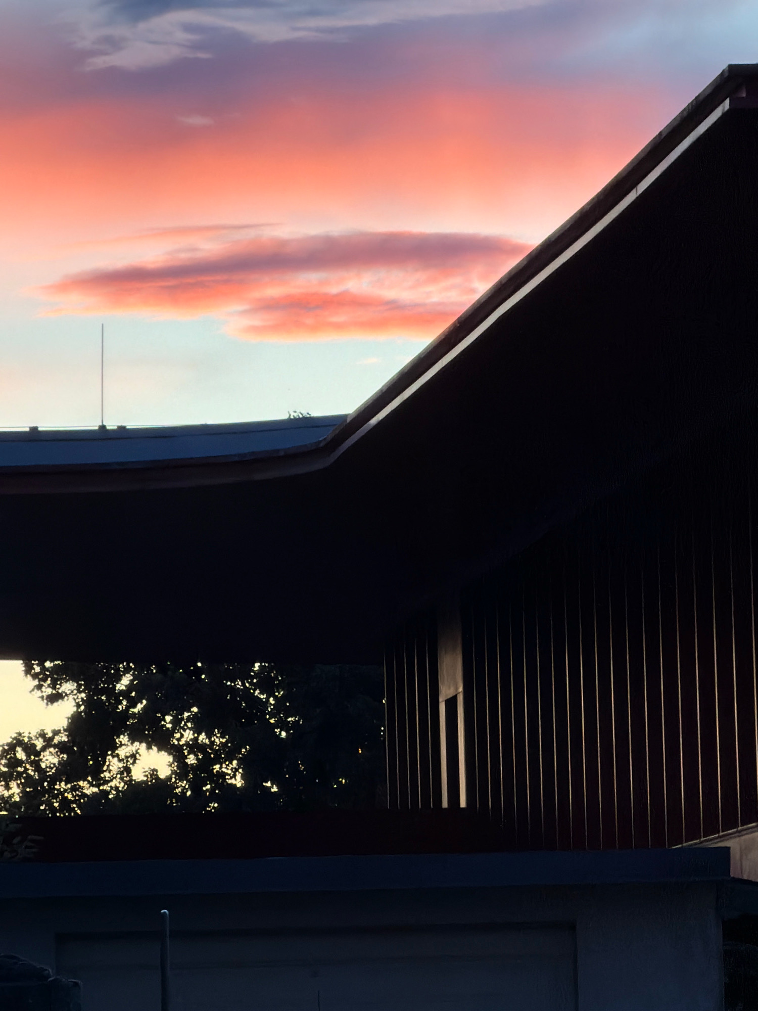 The image shows a section of a modern building with a dark, sloped roof and a vertical ribbed facade, silhouetted against a colorful twilight sky. The sky displays a gradient of colors, from deep blue at the top to vibrant pink and orange hues in the lower portion, with scattered clouds. In the foreground, a lower structure of the building is visible, with a dark horizontal band and a lighter horizontal surface above a concrete wall. The scene appears to be an exterior view of architecture, likely in an urban or suburban setting, given the presence of the building and the hints of trees visible through an opening in the structure. The overall atmosphere is serene, with the dramatic sky providing a striking contrast to the dark, geometric forms of the building. No people or specific actions are depicted. The location is identified as Esch-sur-Alzette, Luxembourg.