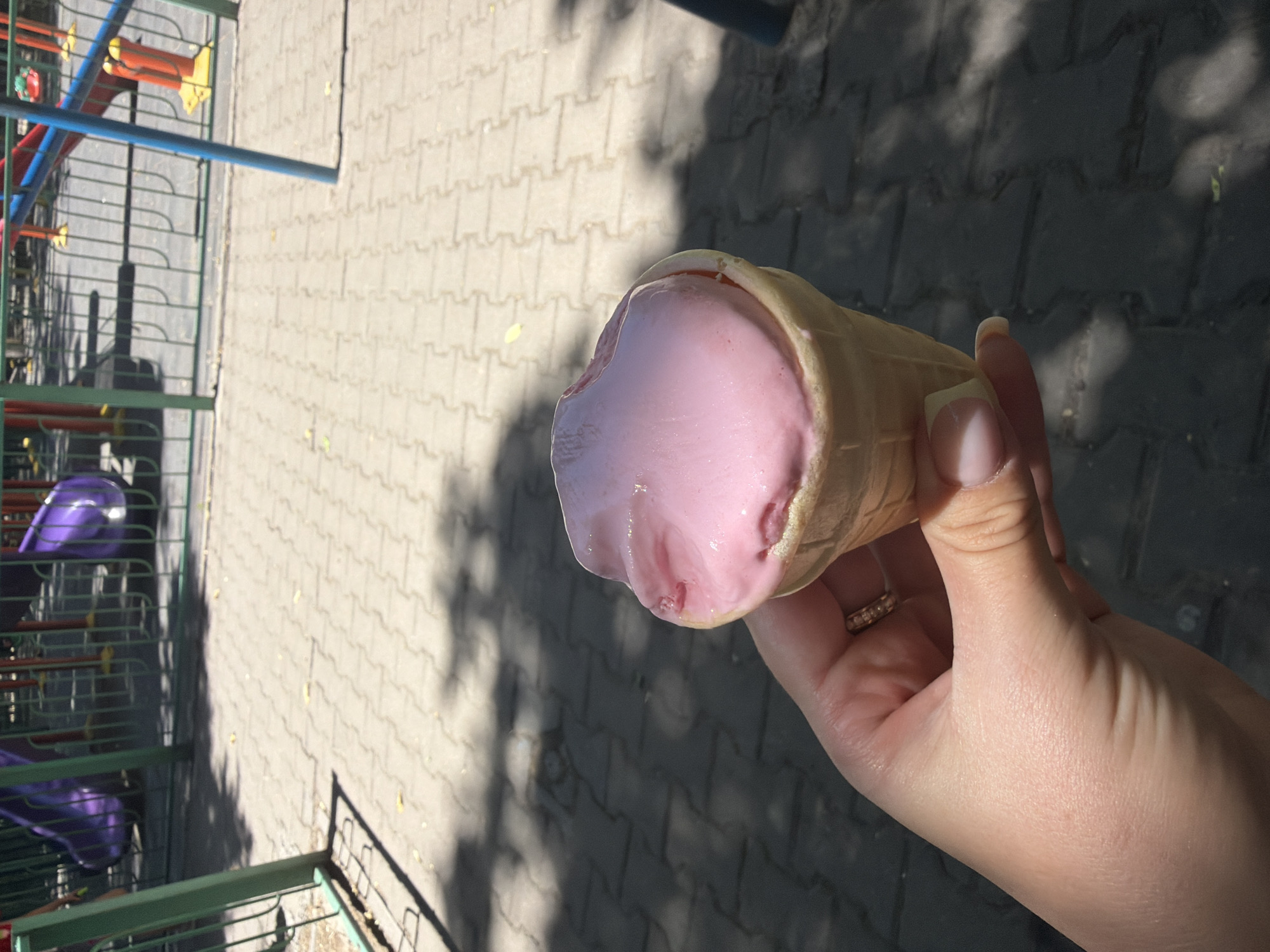 A hand holds a strawberry ice cream cone in a waffle cone. The ice cream is pink and appears slightly melted. The hand has a ring with small stones on the ring finger. The background shows a playground with purple slides and green fencing, and a paved area with interlocking stones. The scene is outdoors and sunny, with shadows cast on the ground. The image was taken in the city of Shymkent, Kazakhstan.