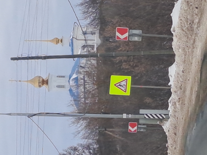 The image displays a winter street scene in Никольское, Russia, under an overcast sky. In the background, two white-walled structures with blue roofs and golden onion domes are partially visible above a line of bare-branched trees or bushes. The larger structure on the left features a prominent golden dome topped with an Orthodox cross. Several dark utility poles, interconnected by overhead wires, occupy the midground. The foreground reveals a road surface and a low bank of dirty, melting snow and slush. Three distinct road signs are present: a central, square, neon-yellow sign with a black border encompassing a white triangle with a black pedestrian figure (indicating a pedestrian crossing); to its left, a small red square sign with a white left-pointing chevron; and to its right, a similar small red square sign with a white right-pointing chevron. A utility pole with black and white diagonal hazard stripes is also visible near the left-side signs. No individuals are depicted.