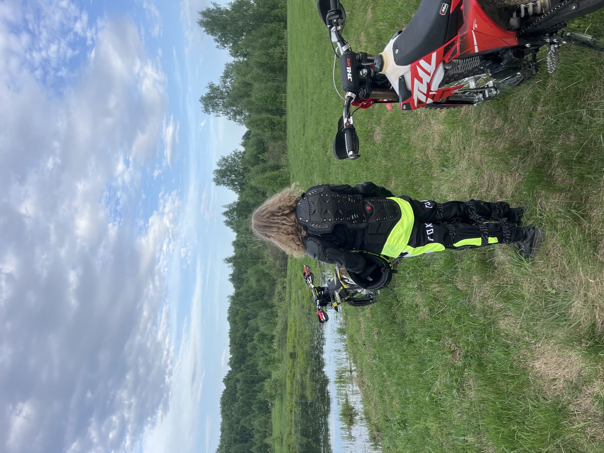 A medium shot depicts a person in motocross gear standing next to a body of water, likely a pond or lake, with two motocross bikes nearby. The person is wearing a black and neon yellow protective suit with "FOX" visible on the pants. The landscape includes green grass and a line of trees in the background. The sky is partly cloudy. Although location details are provided, I cannot confirm that the image was captured in Новочувашский, Russia, or confirm its connection to this place.
