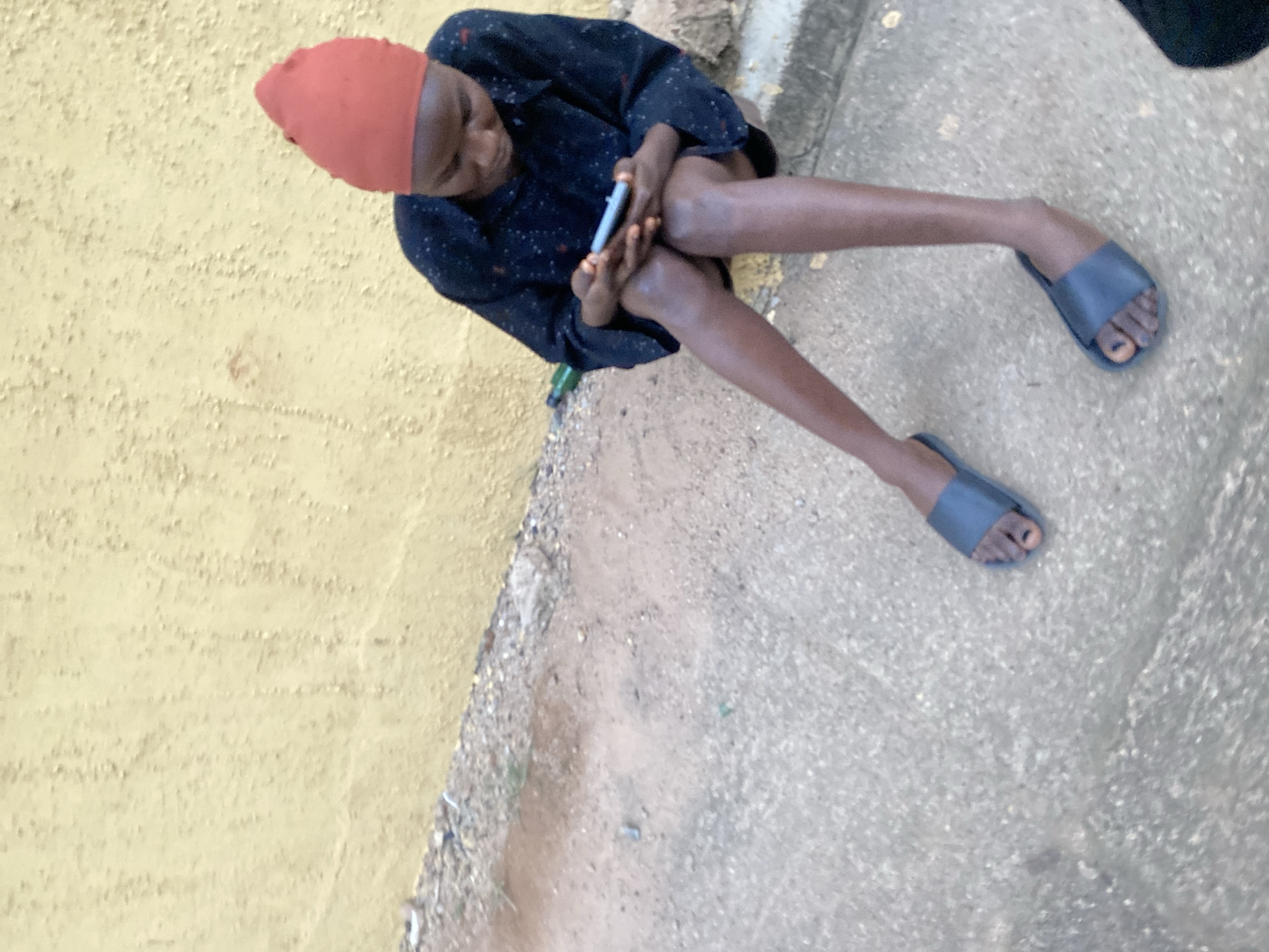 A young person, wearing an orange headscarf and a dark, patterned shirt, sits on the ground, looking down at a mobile phone held in their hands. They are wearing dark, open-toed sandals. The background consists of a rough, textured yellow wall and a dusty, concrete ground with some gravel. A green bottle is visible near the wall. The scene appears to be outdoors, in what could be a residential area or public space. The lighting suggests it is daytime.