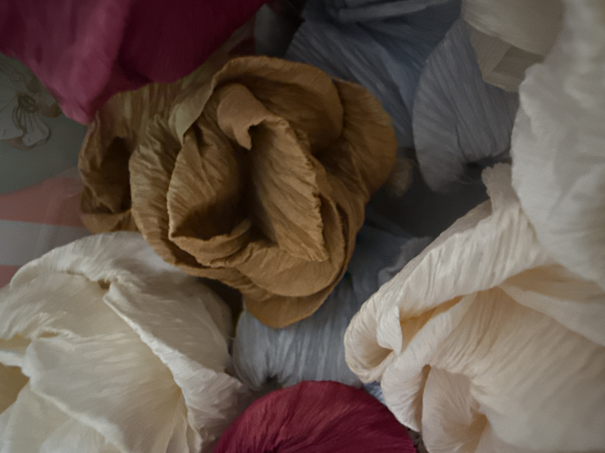 This close-up image presents a vibrant, abstract arrangement of several textured, crumpled objects, likely made of crepe paper or crinkled fabric. The objects are densely packed, overlapping each other, and filling the frame.
The central and most prominent object is a large, brown or tan piece, heavily folded and creased, resembling an abstract flower or a bunched textile. Surrounding it are other similar textured forms in various colors: a large off-white or cream piece occupies the bottom left, while another cream-colored piece is visible on the right. A light blue or grey crinkled object is positioned behind the central brown piece and to the right. Towards the bottom center, a deep red or burgundy textured form is partially visible. In the top left corner, a deep pink or magenta object is shown, behind which a faint, light-colored floral or decorative pattern can be discerned on what appears to be a light green-grey surface, possibly wallpaper or a box lid.
The texture throughout is consistently wrinkled and pleated, creating a sense of depth and softness. There are no people or specific activities depicted. The lighting is soft and diffused, highlighting the folds and contours of the materials. No visible text is present. The scene depicts decorative elements, likely indoors, though no specific location cues for Varna, Bulgaria, are visible within the image itself beyond the objects.