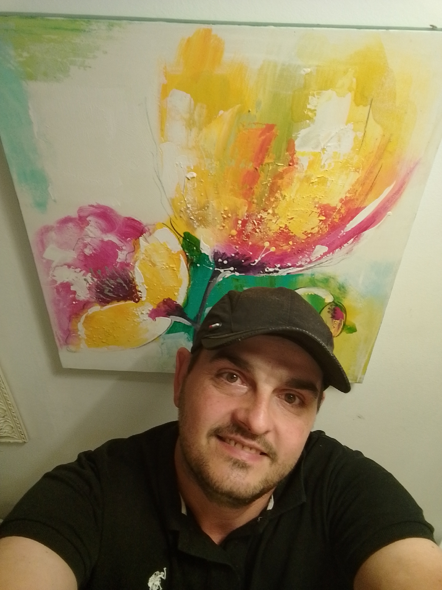 A man wearing a black baseball cap and a black polo shirt is photographed in a close-up, frontal view. He has brown eyes, a short beard, and is smiling. Behind him, a large, abstract painting with vibrant yellow, pink, and green brushstrokes is displayed on a white wall. The lighting appears to be artificial, casting a slight shadow from the man's hat onto his forehead. The environment suggests an indoor setting, possibly a home or gallery.