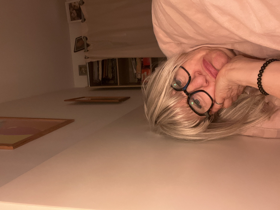 A woman with blond hair and glasses leans down toward the viewer, her cheek resting on her hand. A black beaded bracelet is visible on her wrist. Above her, a pink fabric is draped, possibly a blanket or curtain. Behind, a wall with framed pictures leads to a closet with clothes hanging inside. The scene is dimly lit, with a soft, warm color cast.