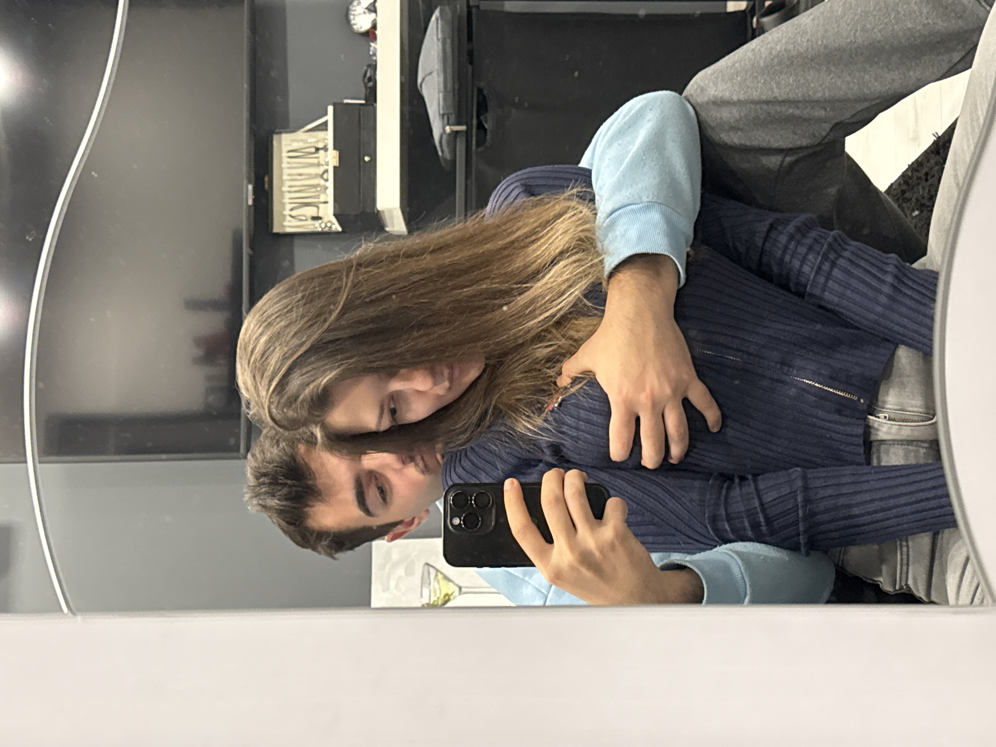A man and a woman are taking a selfie in a mirror. The man is holding the phone, and the woman has her arm around him. They are both wearing sweaters, the man's is dark blue with light blue sleeves and the woman's is a similar color. They are indoors. There is furniture in the background, as well as a picture of a cocktail. It is not possible to determine the location within Sofia, Bulgaria from the given information.