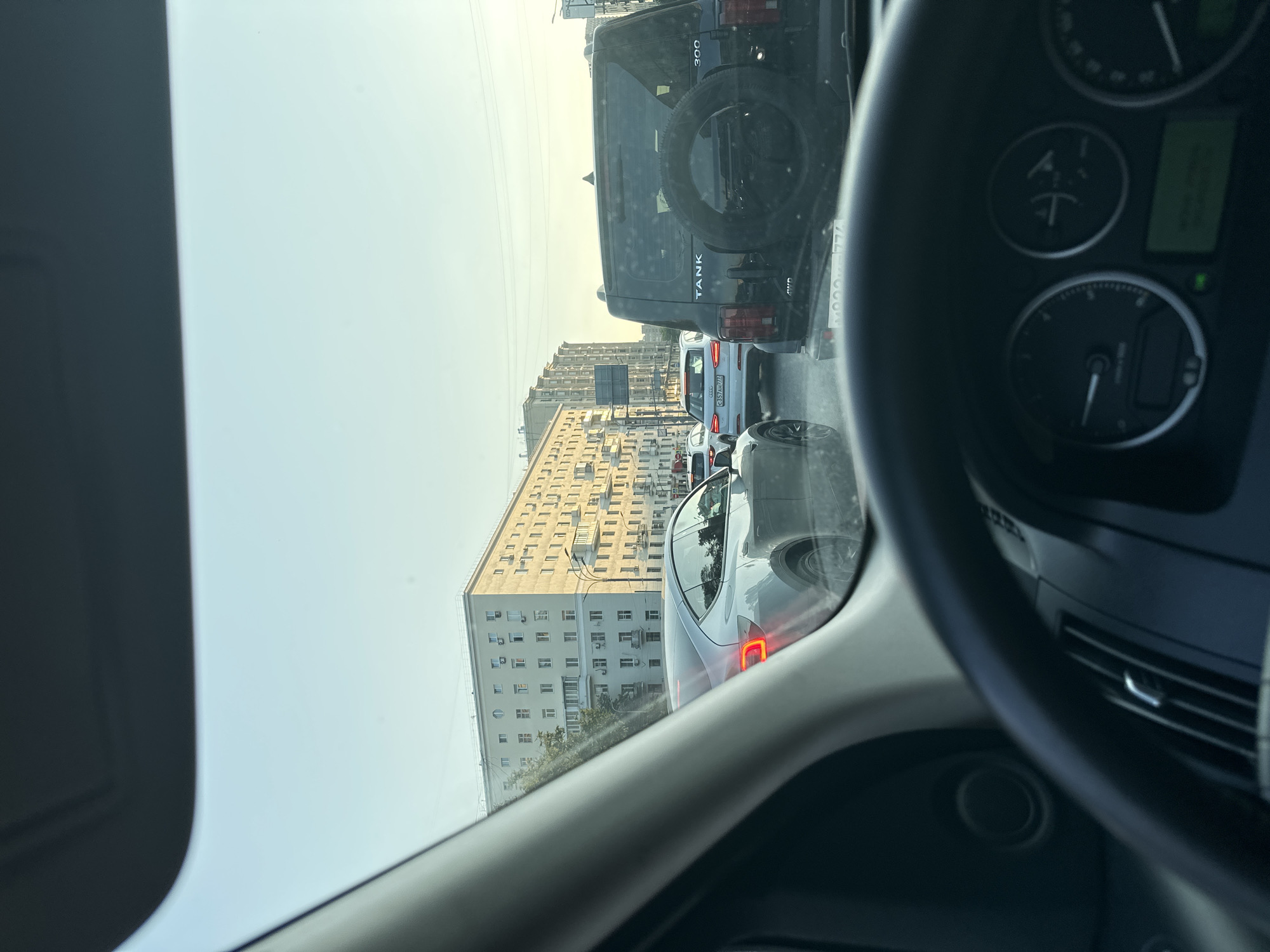 The image depicts a view from inside a car, looking forward through the windshield and side window, likely during a traffic jam. The primary subjects are several vehicles in a line on a road, with a large residential building dominating the background.
In the foreground, a portion of the car's interior is visible, including the steering wheel and dashboard instruments. The steering wheel is dark and the dashboard displays various gauges and a green digital screen.
Outside the car, a black SUV with "TANK" written on its rear is prominent. It has a spare tire mounted on the back. Following this SUV is a white car with a license plate visible, and behind that, other vehicles can be seen. A modern, silver car is directly in front of the camera car.
The scene is set in a city with a tall, light-colored apartment building characterized by numerous windows and balconies, suggesting an urban residential area. The building appears to be an older, Soviet-era style. Trees are visible between the cars and the building.
The sky is bright and appears clear, indicating daytime, likely late morning or afternoon. The lighting suggests it's a sunny day, with some glare on the cars. The presence of traffic and the arrangement of buildings suggest an urban setting, specifically Khimki, Russia, as indicated by the location context. No people are clearly discernible within the vehicles or the scene. The overall mood is neutral, characteristic of a mundane traffic situation.
