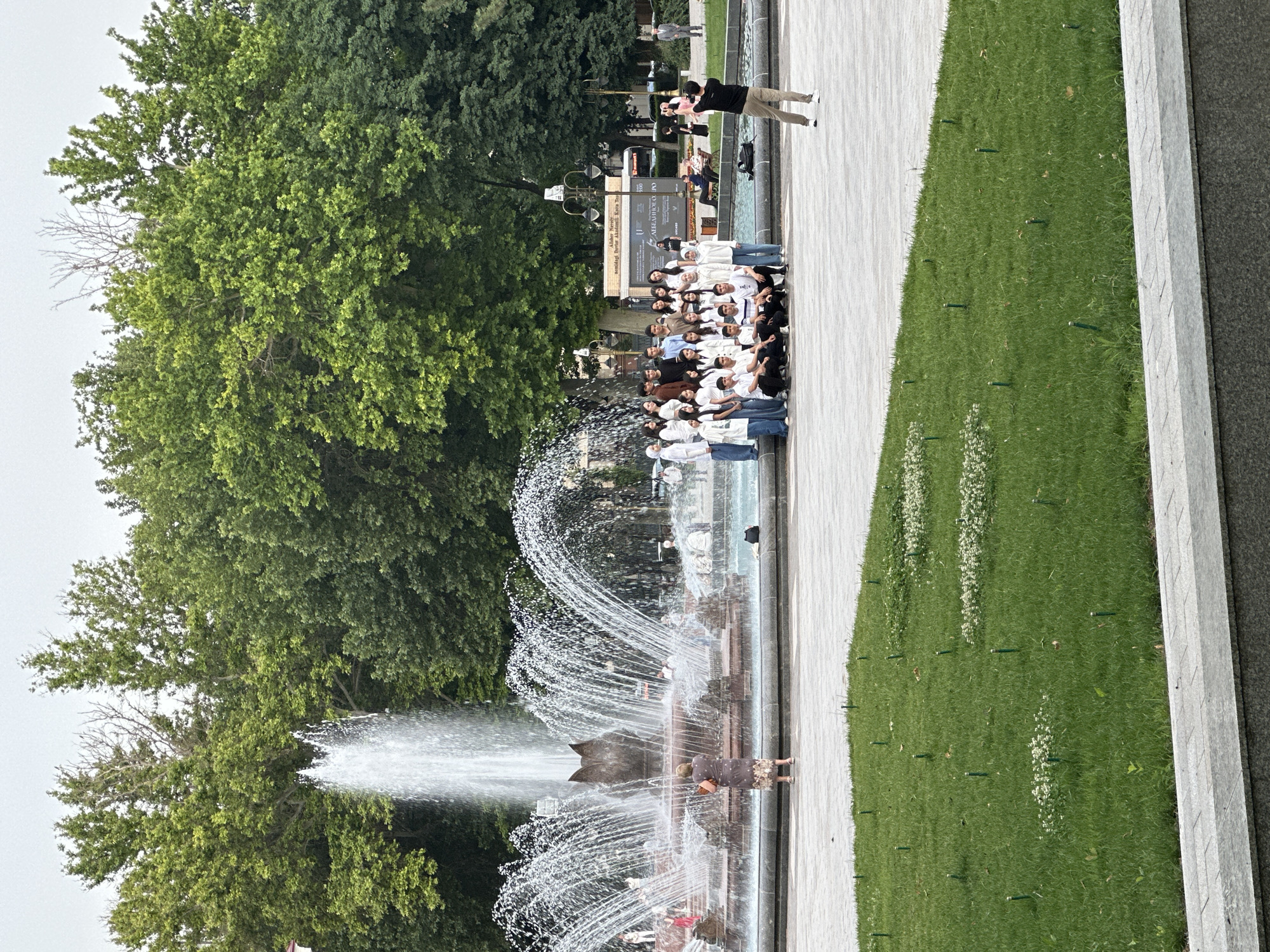 A group of approximately 30 people, mostly young adults, is posing for a photograph near a fountain in Tashkent, Uzbekistan. They are standing close together, some wearing white shirts. A person in dark clothing stands apart, taking a picture of the group. The fountain is multi-tiered with water spraying upwards. A woman is standing near the edge of the fountain. Green trees are visible in the background. A sign with text is visible behind the group. Green grass and a paved walkway are also present.