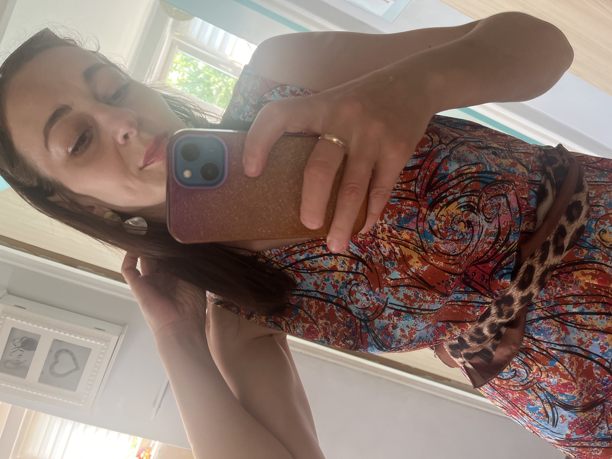 A woman is taking a selfie in a room, reflected in a mirror. She is wearing a colorful, abstract-patterned dress with a leopard print belt. She has long brown hair and is wearing earrings and a ring. Her left arm is raised, and she is holding her phone to take the picture. The background shows a window with blinds and part of a white cabinet with heart decorations. The lighting suggests it is daytime.