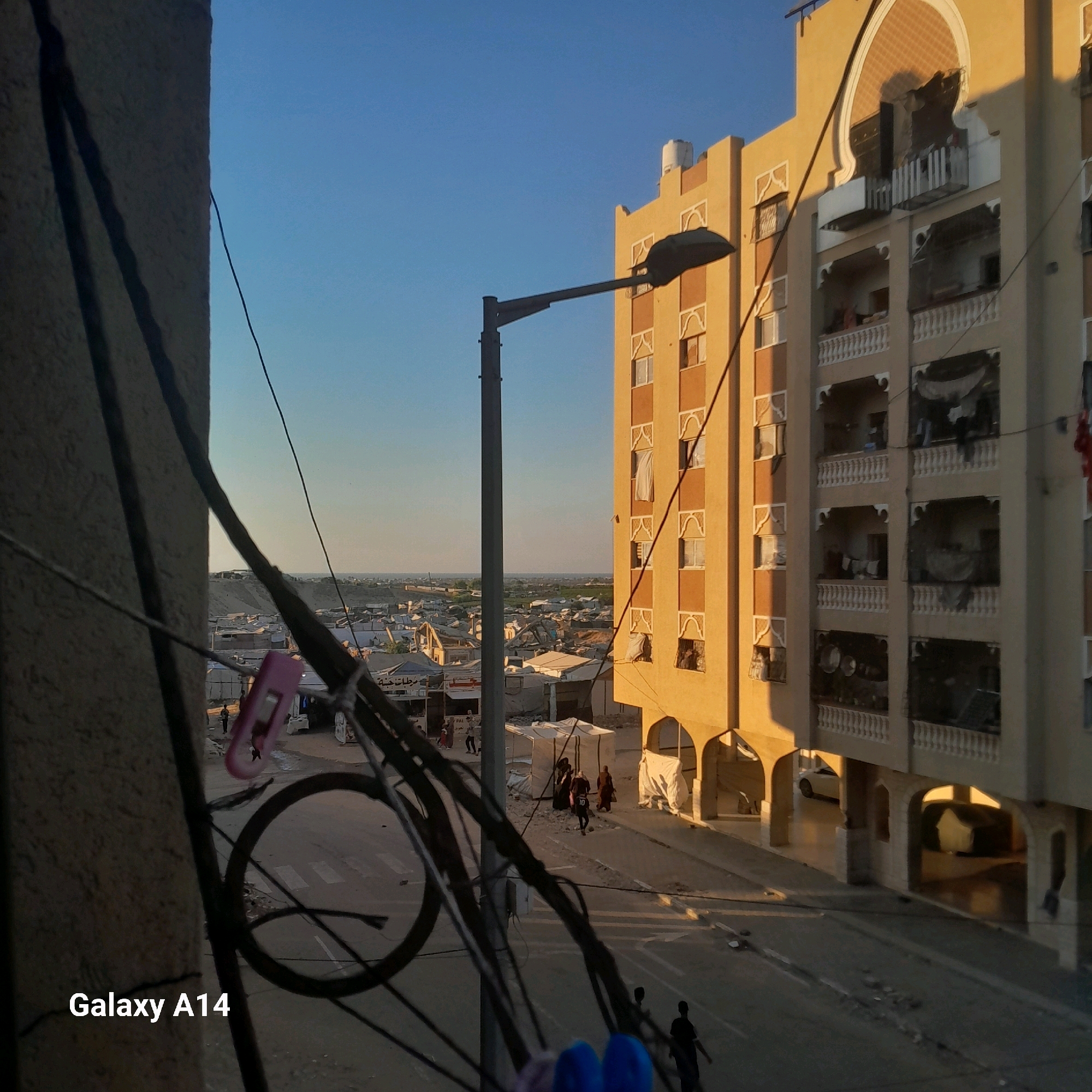 The image, taken in Khan Yunis, Palestinian Territories, captures an urban scene with a focus on a multi-story building and its surroundings. The building, painted in warm, sandy tones, features multiple balconies with ornate white railings and arched openings. Some balconies appear to have makeshift coverings or tarps, while others show signs of habitation with visible objects inside. A lamppost extends over the street in front of the building. The street itself is paved, with some debris scattered on its surface. In the background, a densely populated area with numerous smaller structures, possibly informal settlements or damaged buildings, stretches towards the horizon under a clear blue sky. Several individuals, including women in headscarves and men, are walking on the street and near the buildings. A white car is visible parked under the arches of the building. The foreground is dominated by a cluster of dark electrical wires, casting shadows and obscuring parts of the view, with a pink clip attached to one of the wires. The lighting suggests it is either early morning or late afternoon, with the warm glow of the sun illuminating the building. The overall impression is one of a community in a state of ongoing development or recovery, set against a backdrop of residential and informal structures.