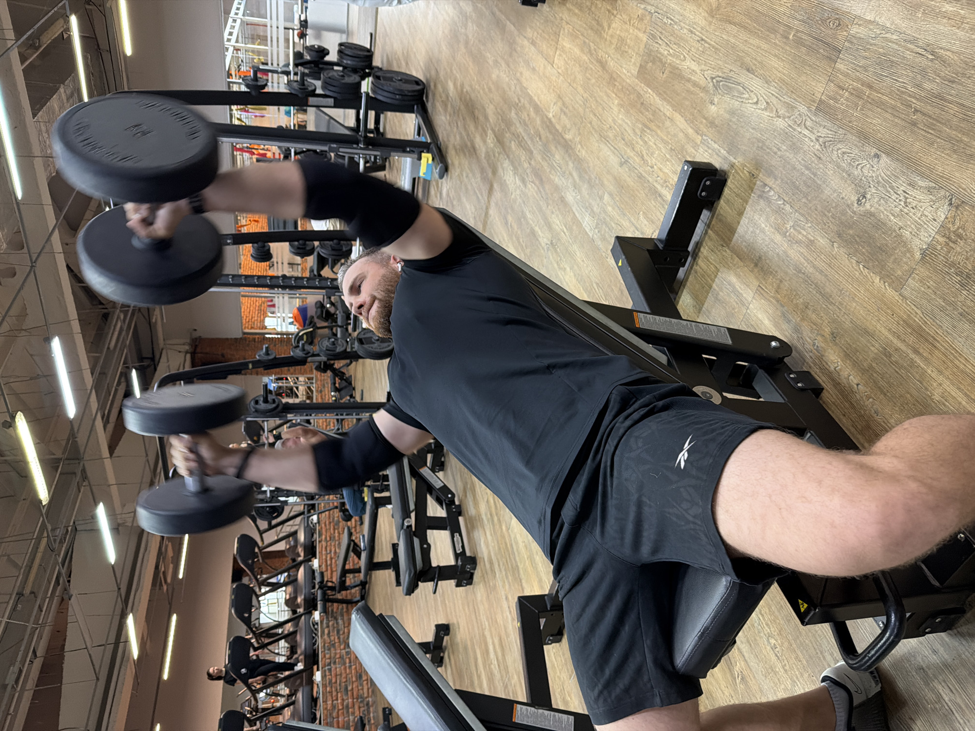 The image captures a man engaged in a dumbbell workout at a well-lit, modern gym. He is lying on a black weight bench, performing an exercise with two black dumbbells, one in each hand. The dumbbells are in motion, showing a slight blur, particularly on the left side of the frame.
The man has a beard and short, light-colored hair. He is dressed in a black t-shirt, black shorts, and black elbow sleeves or compression armbands. A white Reebok logo is visible on the left thigh of his shorts. He appears focused on his exercise, with an earbud in his left ear.
The gym features a light-colored wooden floor and bright overhead fluorescent lighting. In the background, various other gym equipment, including weight racks with numerous black weight plates, exercise machines, and benches, are visible. Some exposed brick walls can also be seen, contributing to the gym's aesthetic. Other individuals are present in the distant background, also working out. Yellow warning labels are partially visible on the gym equipment, but their text is unreadable. The overall setting suggests an active and dedicated fitness environment. The location is a gym, potentially in Chaikovsky, Russia.