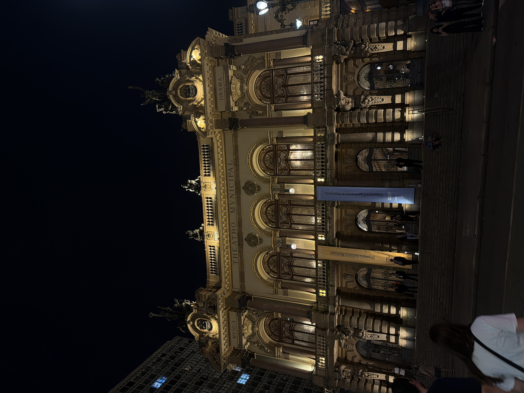 The image shows the Municipal Theater of São Paulo at night. The grand, illuminated building features neoclassical architecture with ornate facades, statues, and large arched windows. Several people are visible on the steps leading up to the theater and on the sidewalk in the foreground. A building with many windows is partially visible to the left of the theater. Two banners hang from the facade of the theater. The street in front of the theater is dark, with the exception of streetlights and the lights from the building itself.