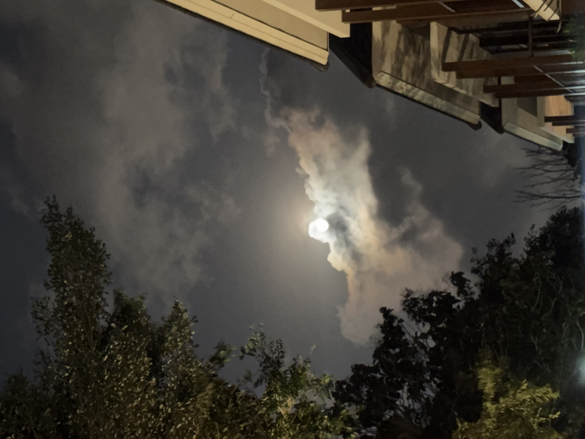 This is a nighttime, eye-level, vertical shot of the sky over Kuta Selatan, Indonesia. A full moon is partially obscured by clouds, casting a bright glow. The branches of a large tree dominate the left side of the frame, and the roofline of a building with wooden pillars is visible on the right. The scene appears tranquil and quiet.