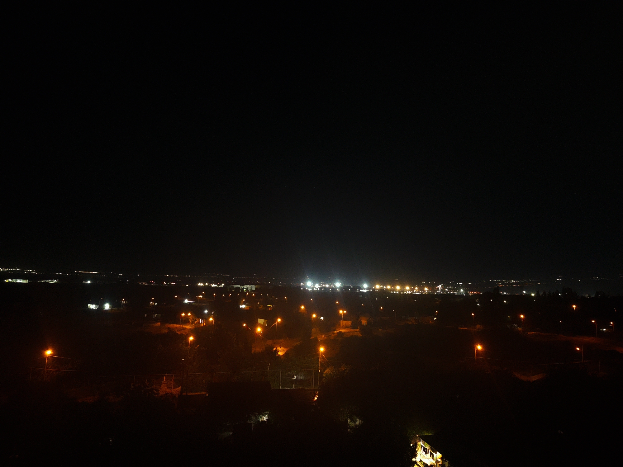 The image is a nighttime aerial view of a city, likely Tbilisi, Georgia, showcasing a landscape dotted with numerous illuminated streetlights and buildings against a dark sky. In the foreground, several streetlights with orange-yellow glows punctuate the lower half of the frame, casting warm light on the surrounding dark terrain, which appears to be partially wooded. Beyond this, the mid-ground reveals more densely packed lights, suggesting residential or commercial areas. The background features a distant city skyline with a concentration of brighter, white lights, possibly indicating a more developed urban center or industrial zone. The overall impression is of a sprawling cityscape at night, with the lights creating a pattern of human activity against the natural darkness of the night sky.