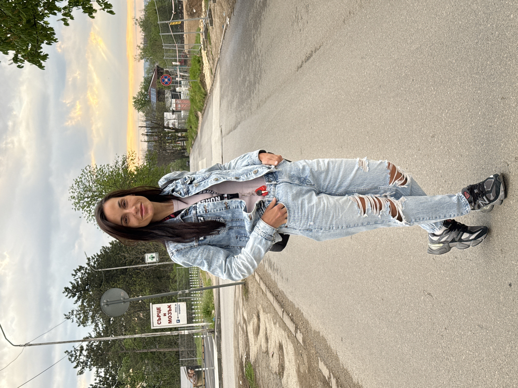 A young woman is standing on a paved road in Pleven, Bulgaria. She is wearing ripped light blue jeans, a light pink t-shirt, a light blue denim jacket, and black and gray sneakers. She has long, dark brown hair and is looking at the camera with a slight smile. The background includes a street with various signs and buildings. One sign reads "СЪРЦЕ И МОЗЪК" (Heart and Brain) with additional text below. Trees and a sky with sunset colors are visible. Construction or renovation activities are also apparent in the background.