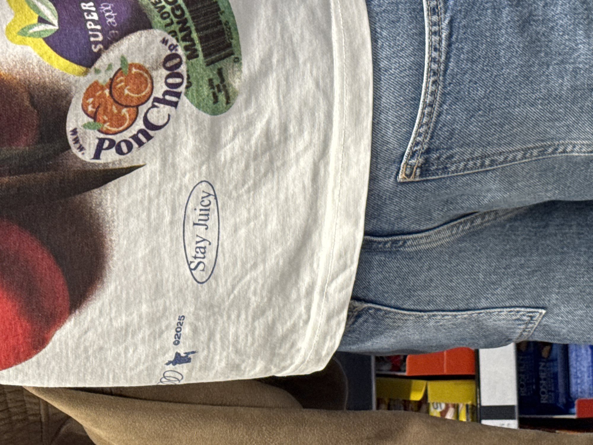 The image shows a close-up of a person wearing blue jeans and a white t-shirt with fruit-themed graphic design. The shirt features images resembling fruit labels with text like "Ponchoo" and "Stay Juicy" with a copyright date of 2025. Below the jeans, boxes are visible which might indicate being in a grocery store.
Without more information about location (address, GPS coordinates) I can not determine with any accuracy if this photo was taken in Sofia, Bulgaria.