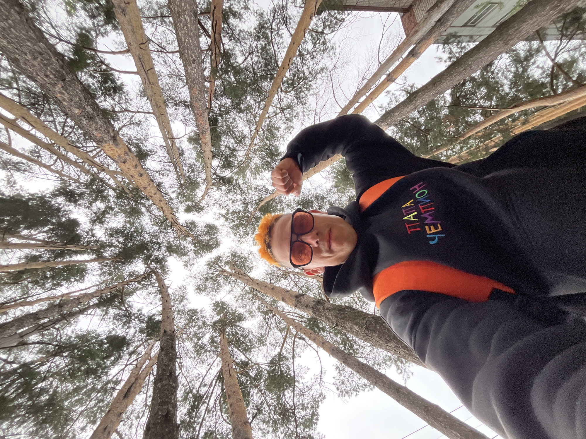 A low-angle, wide shot captures a person with bright orange hair and orange-tinted sunglasses taking a selfie in a forest. The person wears a black hoodie with an orange backpack visible and is flexing their bicep. The hoodie has colorful text embroidery. The background features tall pine trees, their branches reaching upwards against an overcast sky. To the right of the trees, the upper floors of a light-colored brick building with windows are visible, suggesting the location is on the outskirts of a city or town. This scene is consistent with the environment found in the city of Chaikovsky, Russia, known for its surrounding natural areas and urban development.