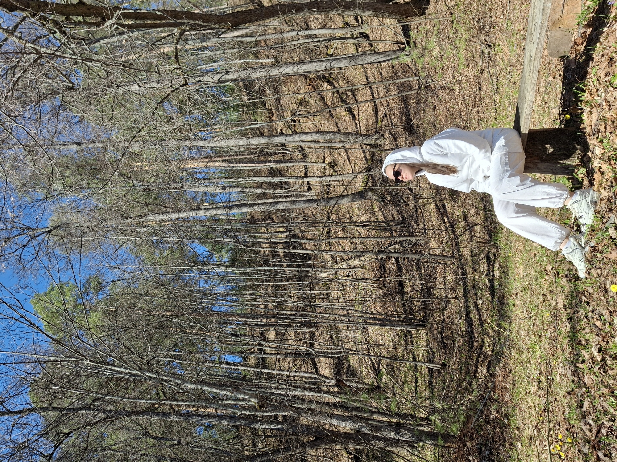 A medium shot captures a person in a white hooded tracksuit and sunglasses sitting on a log in a wooded area. The environment consists of tall, slender trees, many bare, with a mix of brown leaves and sparse green vegetation on the forest floor. The sky is visible in patches through the tree branches. There is no indication of the image being in Noviy, Russia.