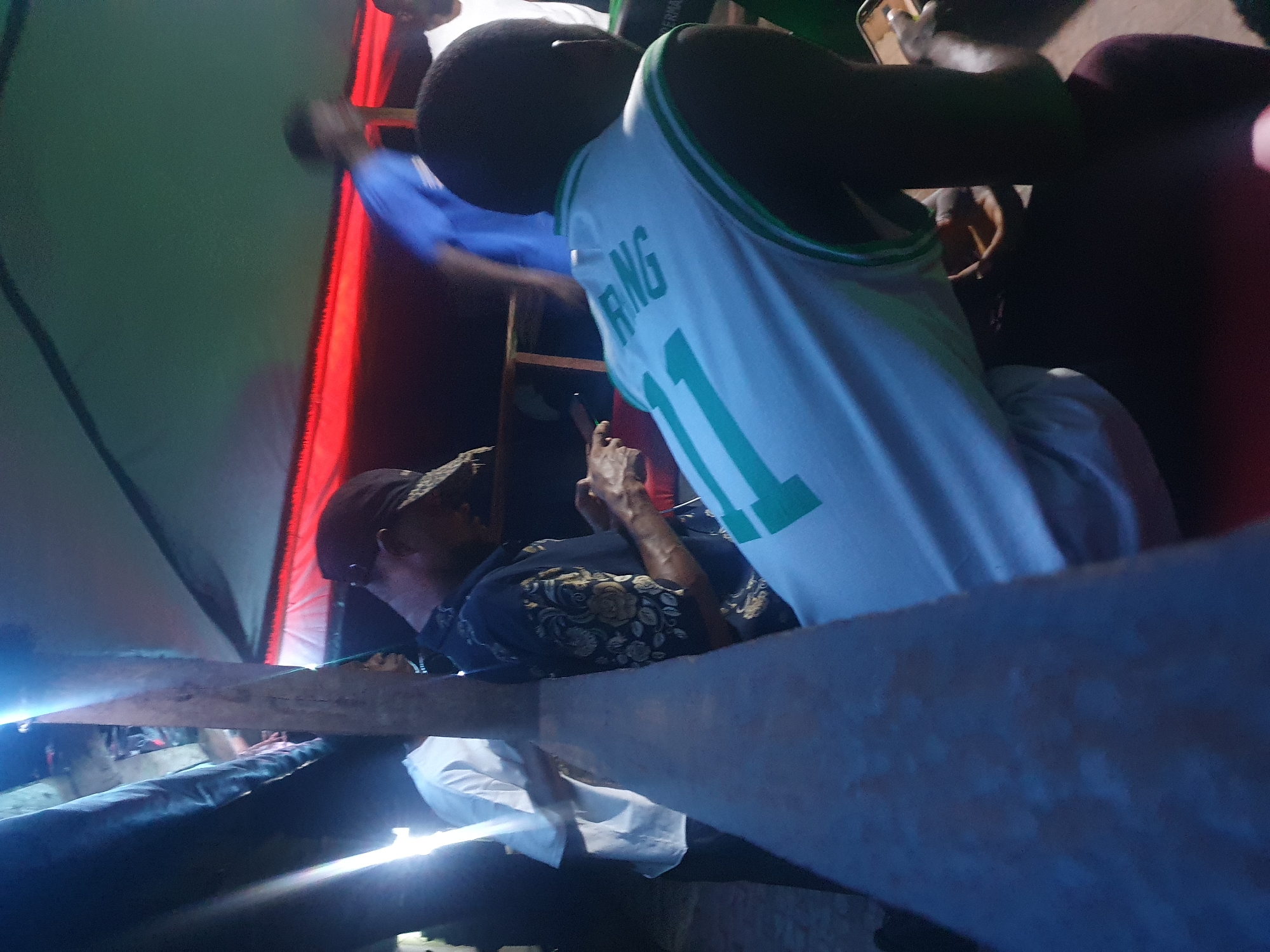 The image, taken in Lokoja, Nigeria, captures a dimly lit scene from a slightly elevated perspective. In the foreground, with their back to the viewer, is a person wearing a white sleeveless jersey with green trim. The jersey prominently features the word "RING" above the number "11" in green lettering.
To the left of this person, another individual is visible, wearing a dark, patterned short-sleeved shirt and a dark cap. This person is engrossed in their smartphone, held in their hands. Further back and to the left, a person in a blue shirt is partially seen, and another person in a white shirt is visible behind a wooden structure.
Two prominent wooden beams run horizontally across the lower half of the frame, possibly acting as a barrier or part of the venue's construction. The background consists of draped fabrics, with a large section of green visible on the left and a bright red section on the right, suggesting a tent, canopy, or internal partitioning.
The lighting is low, indicating an evening or nighttime setting, with several bright light sources casting strong glares and reflections, particularly on the wooden surfaces. The overall atmosphere appears casual, with individuals engaged in their activities within this informal gathering.
