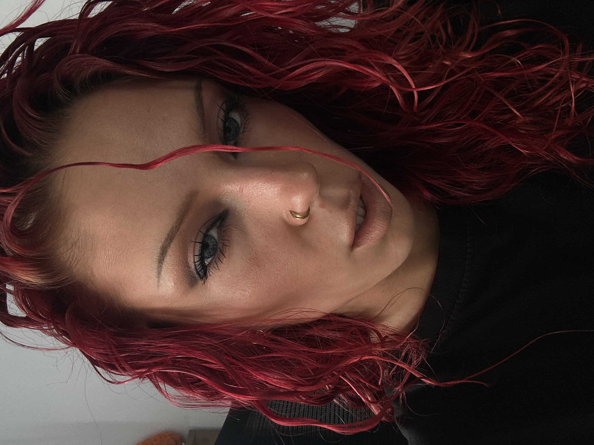 A close-up, eye-level, medium shot captures a woman with vibrant red, wavy hair and a black shirt. She has a nose ring and subtle makeup. The background is indistinct, suggesting an indoor setting. No specific location in North Kuta, Indonesia is identifiable.
