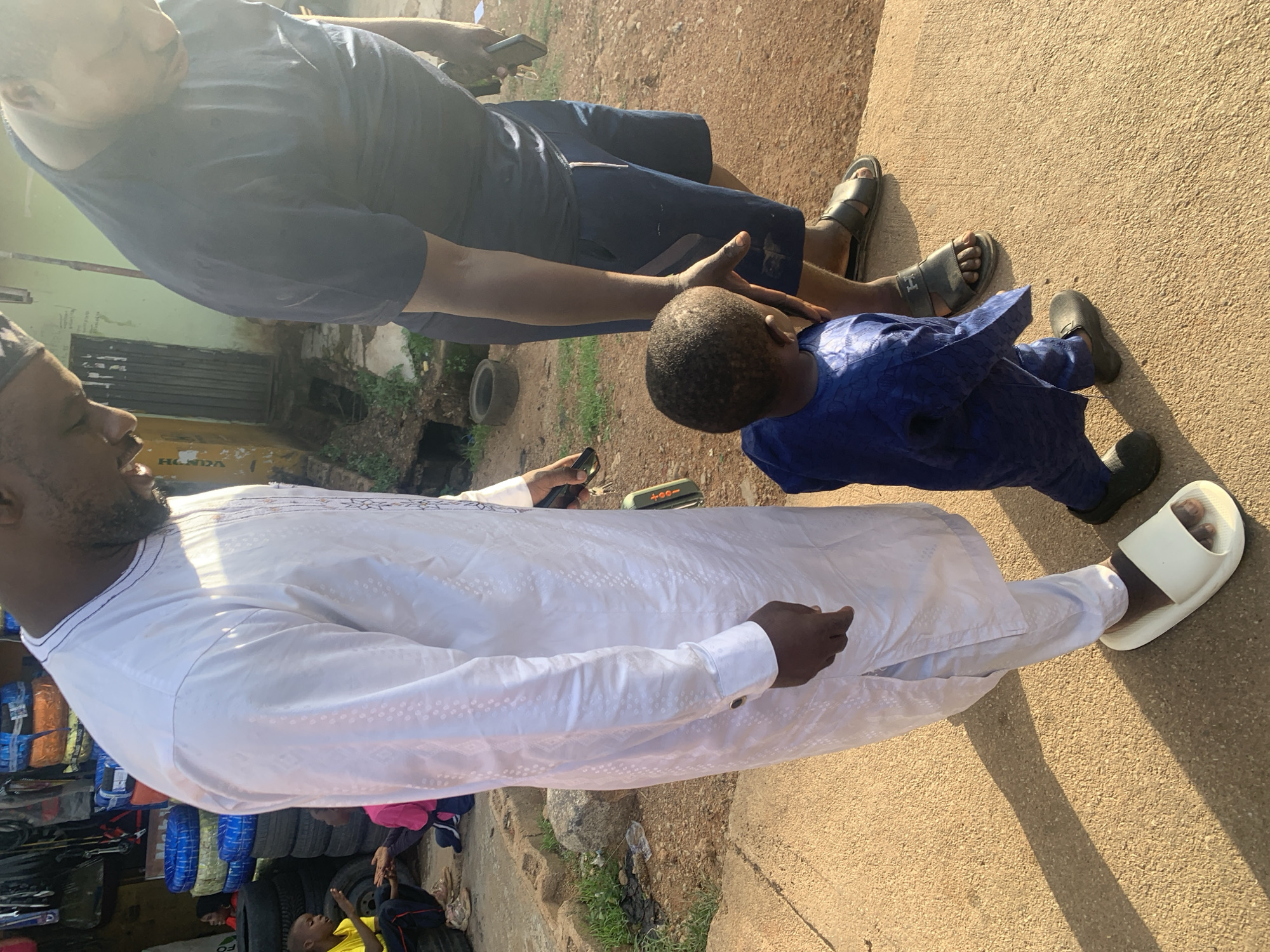 The image depicts a street scene in Jos, Nigeria, likely near a small business or market stall. In the foreground, a man in a white kaftan stands beside a young boy wearing a blue kaftan. The boy is facing away from the camera, looking up at another man, who is partially visible from the shoulders down, wearing a dark blue t-shirt and shorts. The man in the white kaftan is also looking down, possibly at the boy. Behind them, stacked tires and other goods are visible, suggesting a commercial area. The ground is a dusty, reddish-brown earth, and shadows from the figures are cast across it. In the background, there are buildings and a glimpse of green vegetation. The overall impression is a snapshot of everyday life in a Nigerian city.