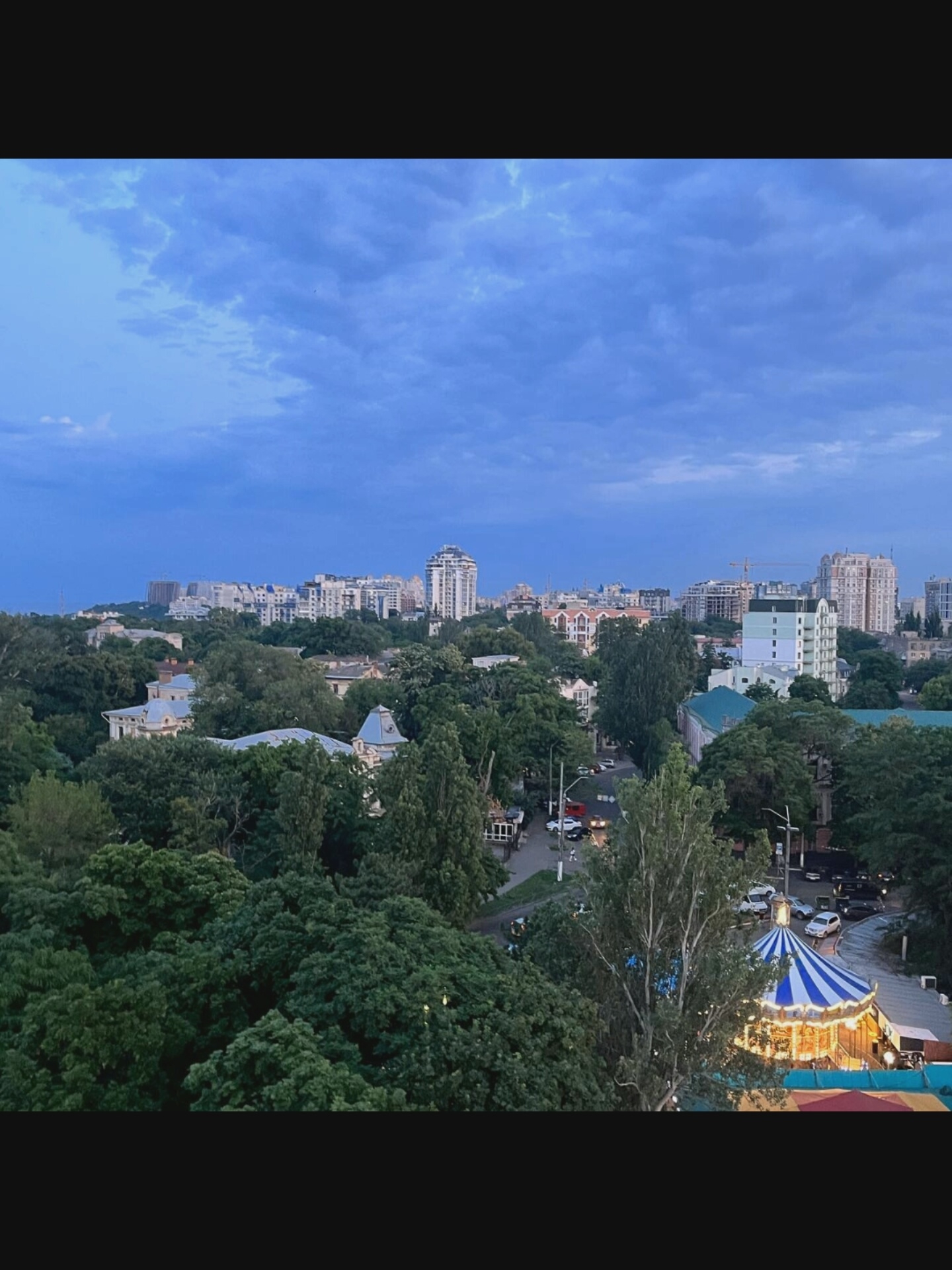 This is an aerial medium shot of a city with a park in the foreground. The sky is a deep blue with a light cloud cover. The city is densely populated with apartment buildings and some construction sites. There is a park with lush green trees in the foreground, with a carousel visible. The carousel is lit up and appears to be operational. Several cars are parked on a street near the carousel. The overall scene suggests an urban environment with recreational amenities. The location is specified as Bielawska Wieś, Poland.