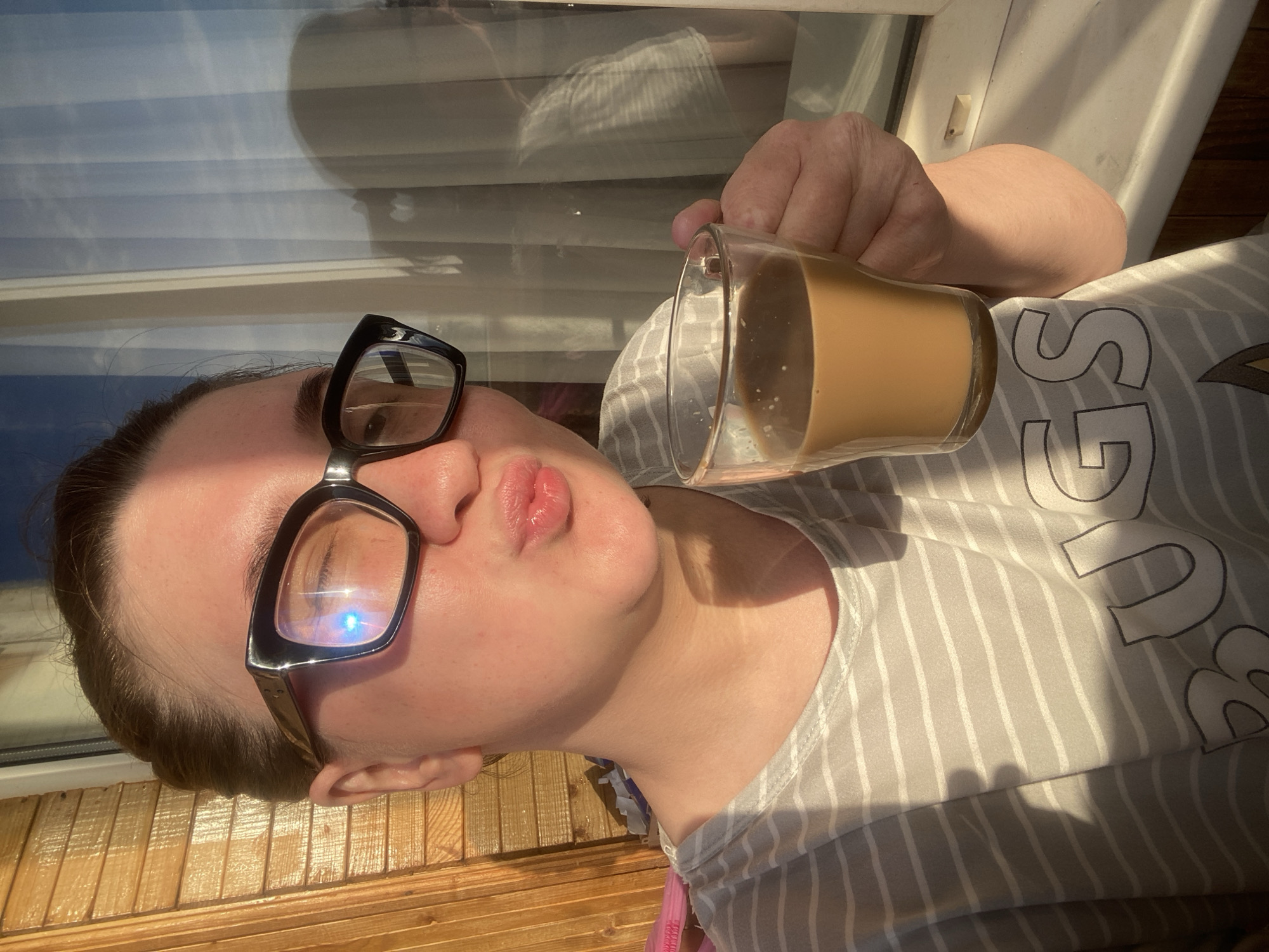 A fair-skinned person with brown hair pulled back is holding a clear glass mug filled with a light brown liquid, possibly coffee or tea with milk, close to their lips as if about to drink. The person is wearing large, black-framed glasses. They are also wearing a gray and white striped shirt with a blurred graphic print on the chest. The background is a window with closed blinds, reflecting outdoor light. Below the window is a wooden surface, possibly a windowsill or porch. The scene appears to be in a domestic setting, likely indoors. The image was likely taken in the city of Chaikovsky, Russia.