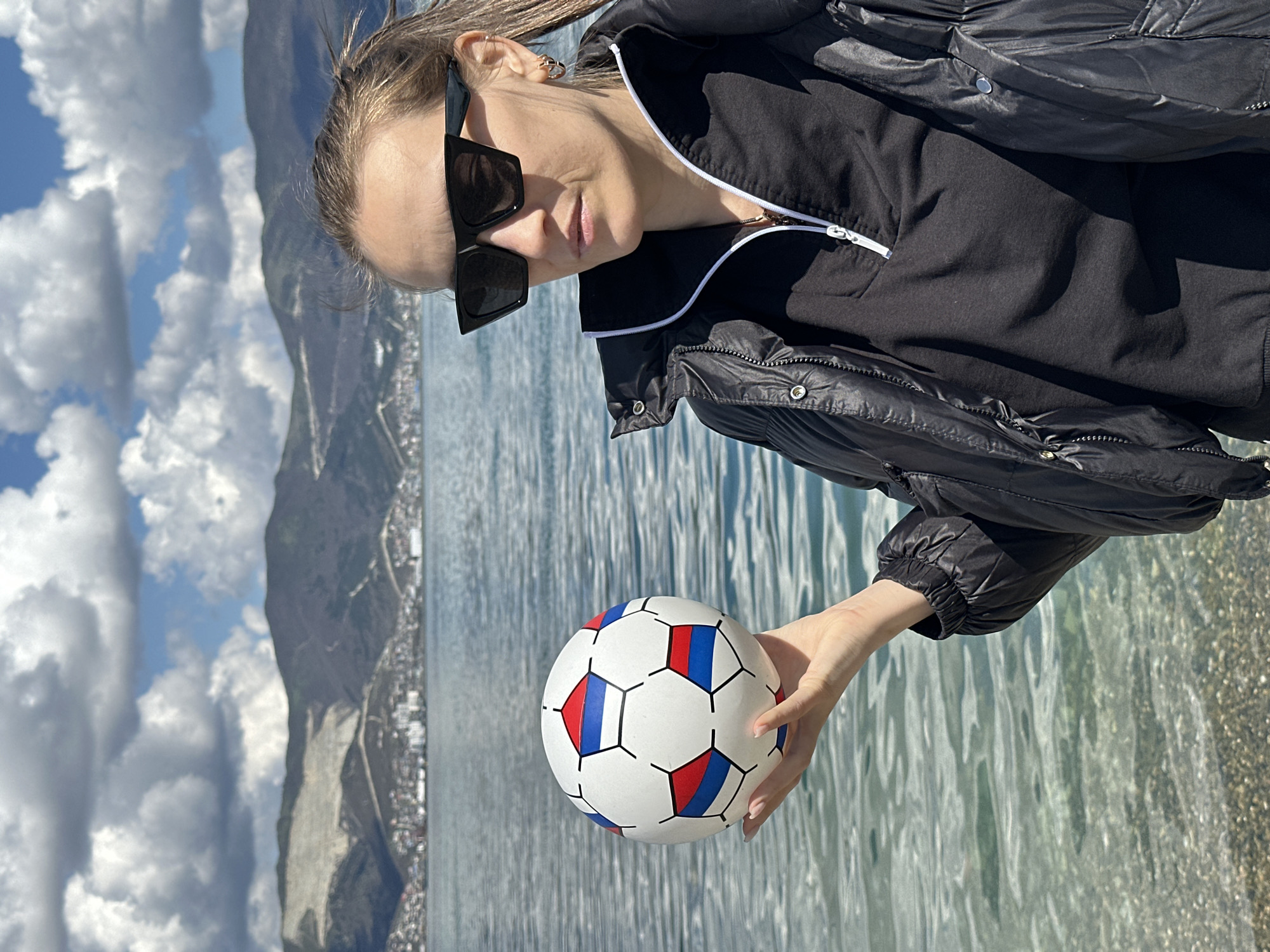 A woman with fair skin and sunglasses holds a soccer ball in front of a body of water. She is wearing a black jacket and a black top with a white zipper. The soccer ball is white with red and blue colored patches. In the background, there are mountains, the sky, and clouds. The location is Myskhako, Russia.