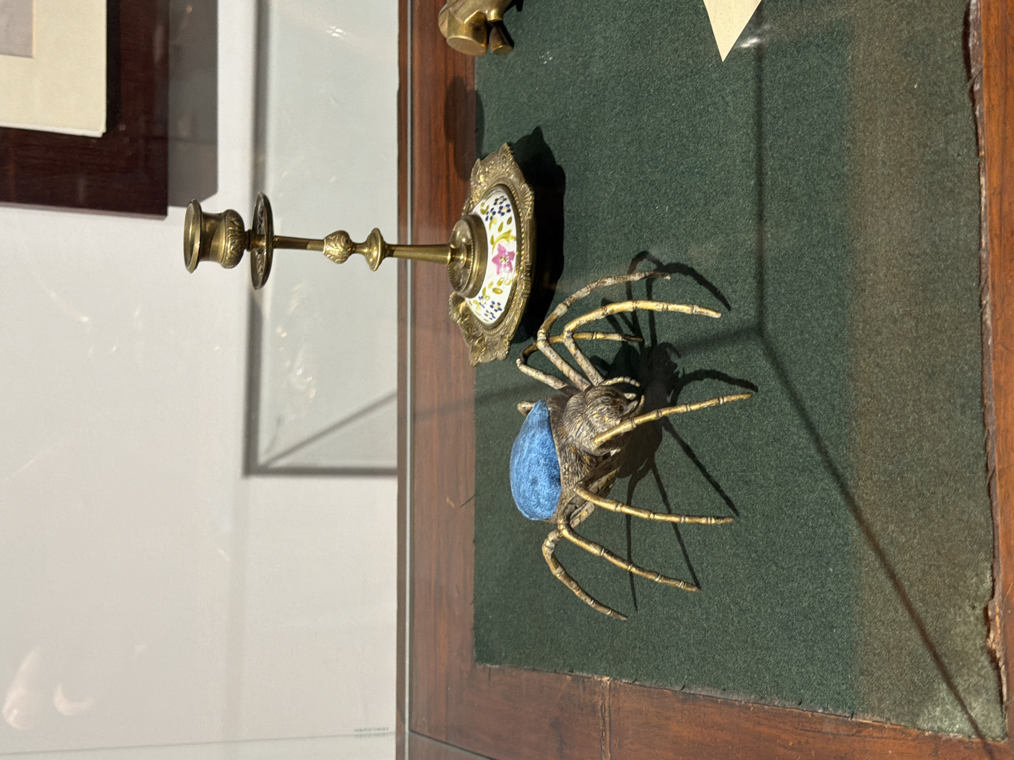 The image depicts a framed display case, possibly in a museum or private collection. The case is made of dark wood and contains several objects on a dark green felt backing. To the left, a gold candlestick with a floral patterned base is visible. Dominating the right side of the frame is a decorative spider figure, likely made of metal with a blue, perhaps velvet or fabric, body. The scene appears to be indoors, with a white or light-colored wall and another framed object visible in the background. There are no people visible in the image. The specific location within Sofia, Bulgaria, cannot be determined from the image alone.