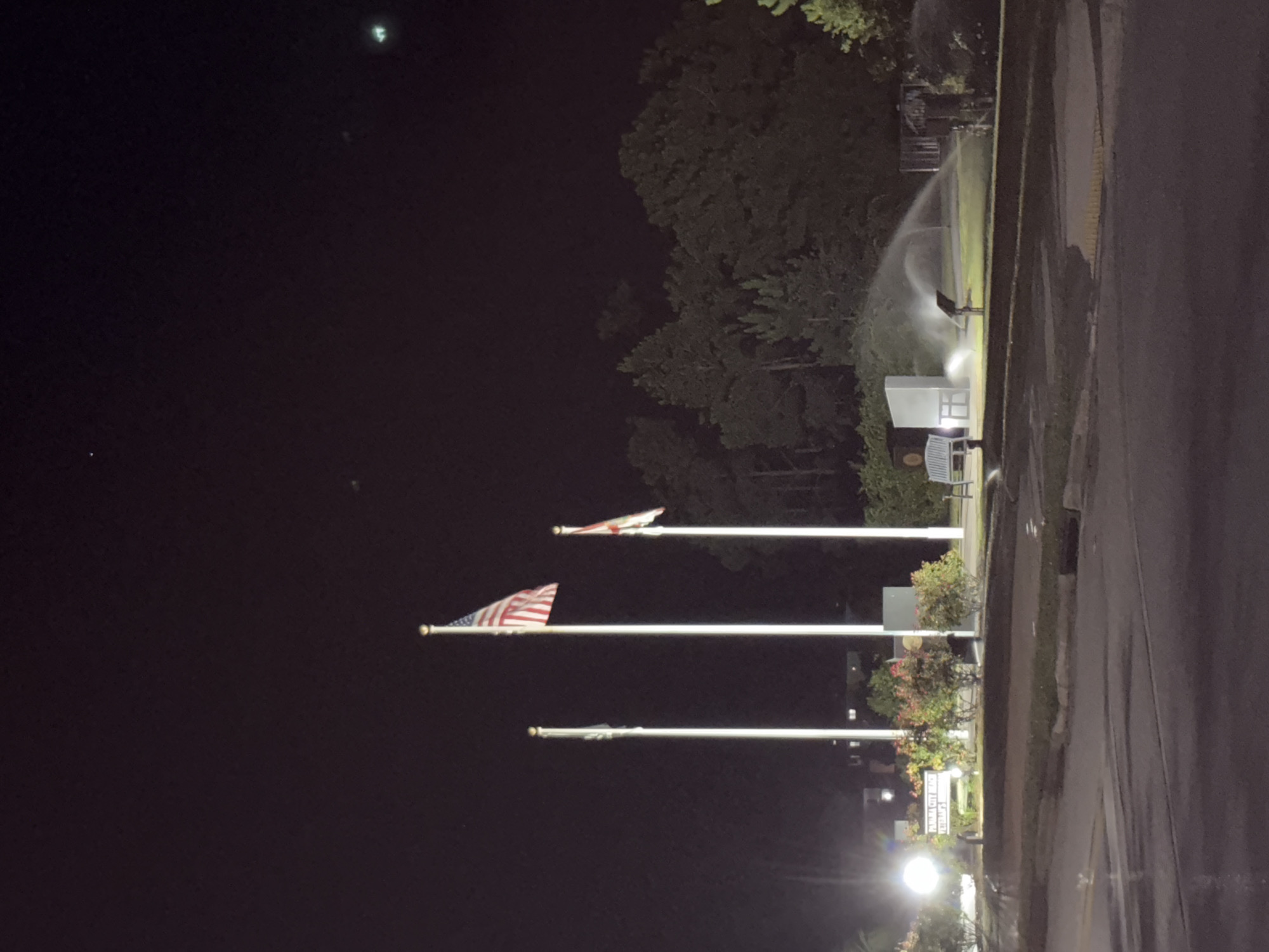 The image shows a night scene in Panama City Beach, Florida, United States of America. In the foreground, a wet asphalt road is visible. Behind it, a grassy area is illuminated by artificial lights. Several flags are planted on poles; one is the American flag, and another appears to be a state flag. A sign with text, possibly for "Panama City Beach Veterans' Park," is illuminated by a bright light. There are also benches and a sprinkler system actively watering the grass. Trees are visible in the background. The overall impression is a memorial or park area at night.