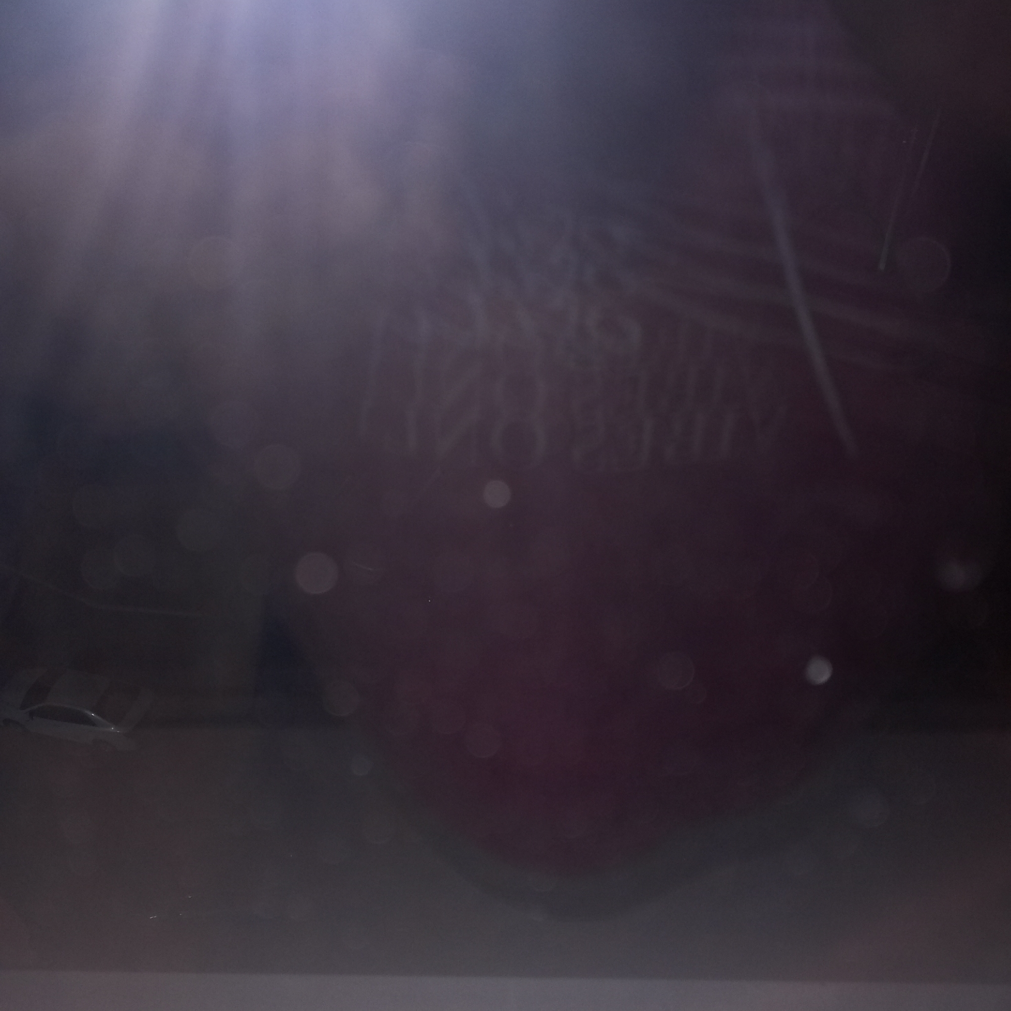 The image is a close-up, poorly lit, and blurry shot of what appears to be a dark red object with some text written on it in white. The text is illegible due to the blurriness and low light conditions. There are also several circular bokeh effects scattered throughout the image, suggesting a shallow depth of field. Additionally, there are light streaks and dust particles visible, indicating a possible lens flare or ambient light. The bottom part of the image shows a lighter, possibly gray surface. The context of Prokopyevsk, Russia, is not deducible from the visual information provided in the image.