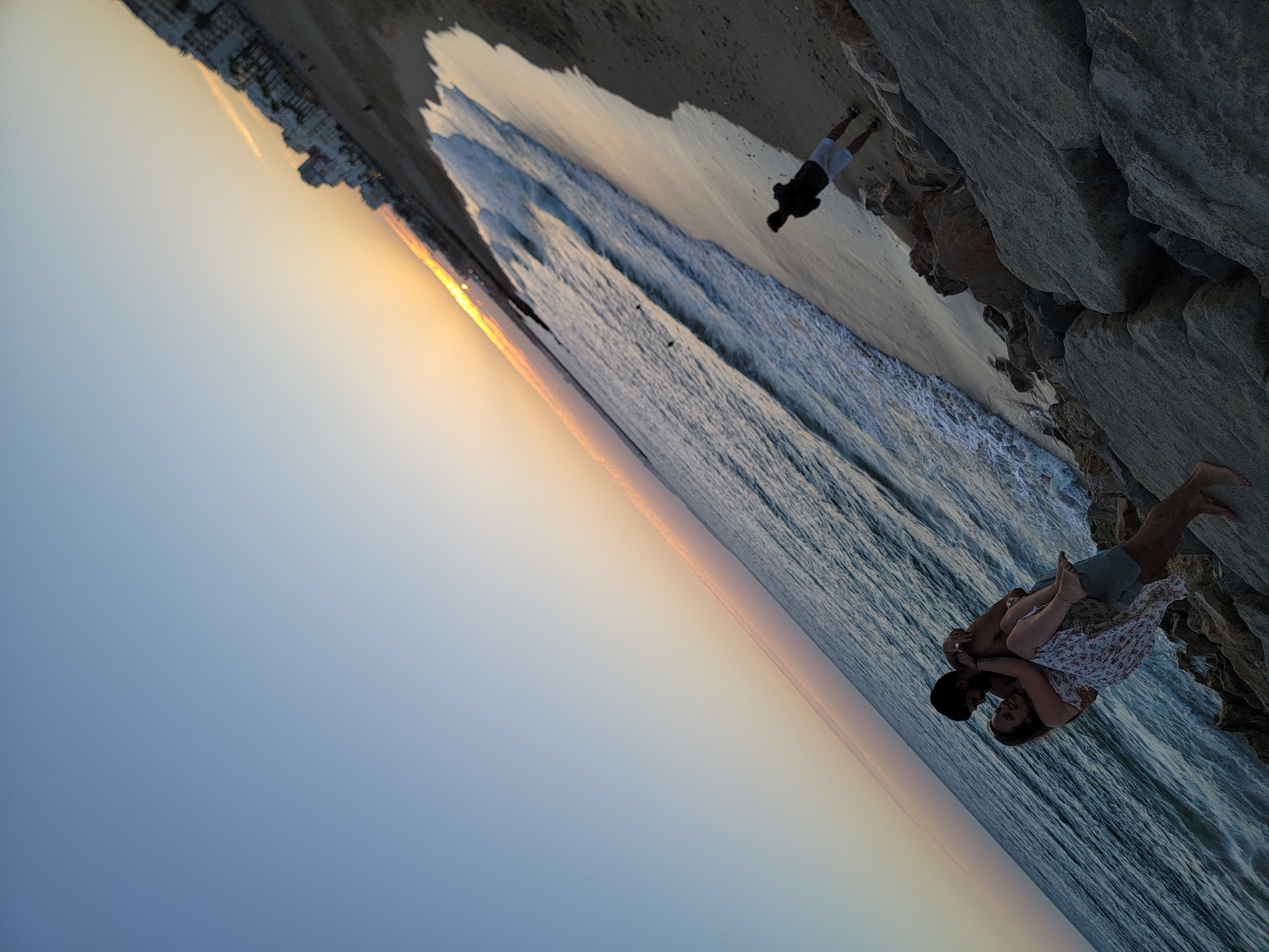 This image captures a romantic scene on a beach in Quarteira, Portugal, at sunset. A man is holding a woman in his arms, both looking at each other and smiling. They are positioned on a rocky shoreline. In the background, the gentle waves of the ocean meet the sandy beach, and a line of buildings is visible along the coast, illuminated by the soft glow of the setting sun. Another person, a man wearing a dark top and light shorts, is standing further down the beach, facing away from the camera. The sky displays a gradient of blue to orange, indicating the late hour. The overall atmosphere is one of intimacy and serene natural beauty.