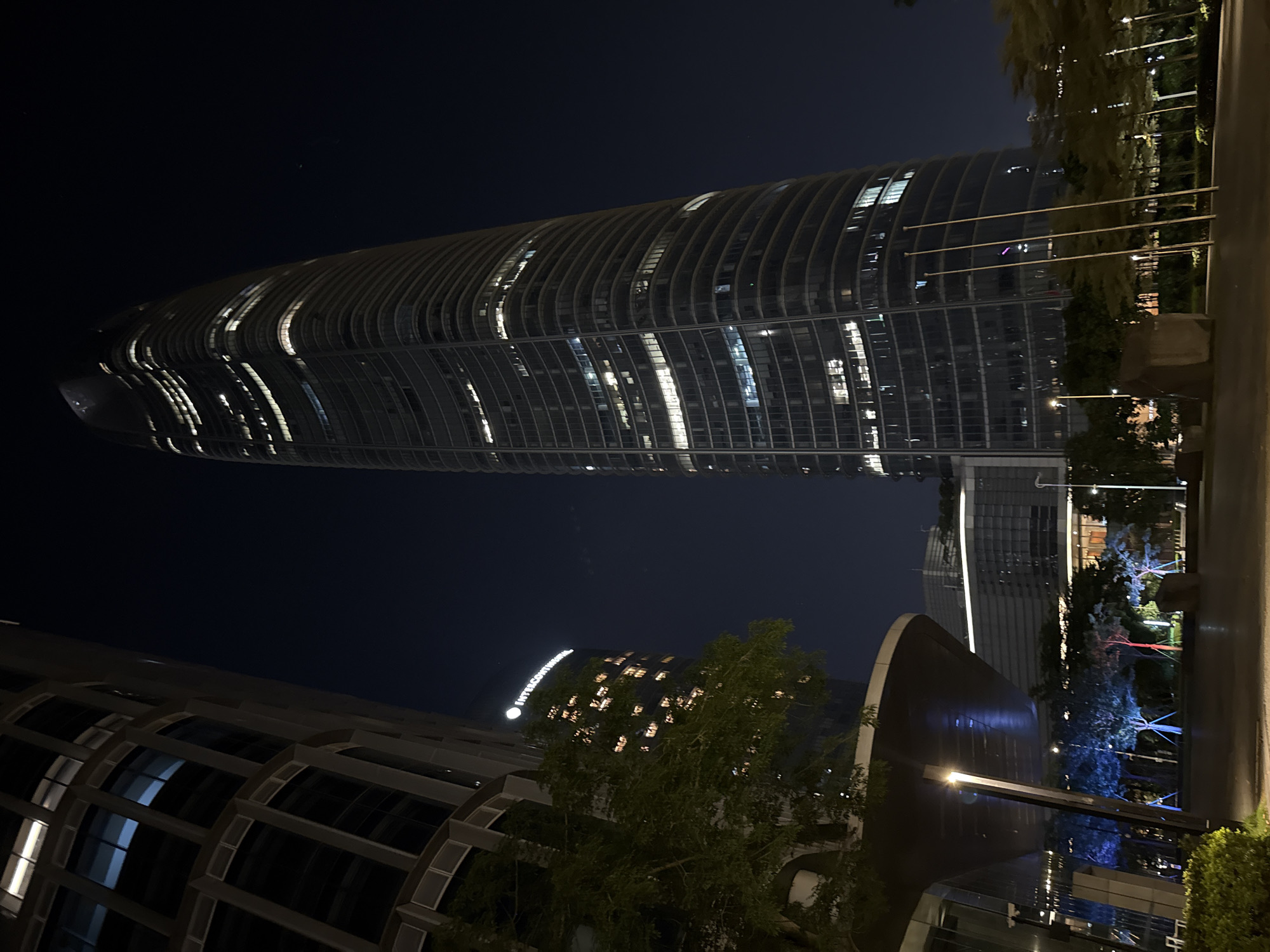 This is a night-time, eye-level, vertical, medium shot of a city skyline in Guangzhou, China. The prominent subject is a large, curved skyscraper with numerous windows illuminated from within. The building's facade appears to be made of glass and metal, with a distinctive ribbed or layered design that creates a sense of verticality and curvature. The dark night sky serves as the backdrop, emphasizing the artificial lights of the city.
In the foreground, to the left and bottom of the frame, parts of other buildings are visible, also with illuminated windows, suggesting a dense urban environment. To the right, a line of trees with their lights on is present, along with flagpoles. The overall setting is an urban landscape at night.
There is some visible text on a building in the middle ground: "INTERCONTINENTAL". This suggests the presence of a hotel or a branded establishment. The time of day is clearly night due to the dark sky and the illuminated buildings and streetlights. The weather appears clear, as there are no visible signs of rain or fog. The atmosphere of the scene is modern and dynamic, typical of a bustling metropolitan area at night. No people are visible in the frame.