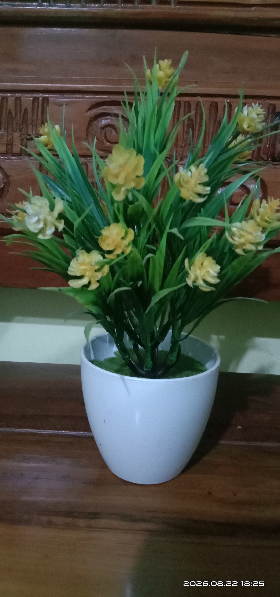 A close-up, eye-level shot features a white pot containing a green, artificial plant with small yellow flowers. The pot is situated on a dark wooden surface. In the background, a section of dark wood furniture with carved decorative patterns is visible. The scene is lit, casting subtle shadows. The timestamp "2026.08.22 18:25" is present in the lower right corner. The location is identified as Pakong, Indonesia.