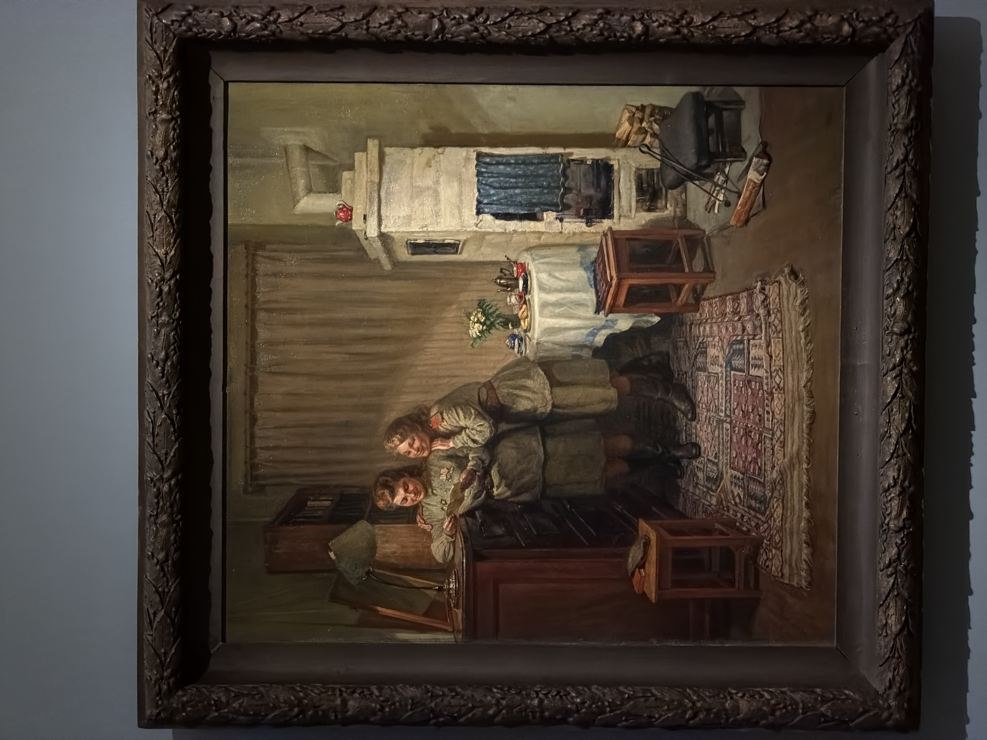 The image is a framed painting depicting an interior scene, likely set in a Russian home. Two young girls, possibly sisters, are the central figures. They are dressed in what appear to be matching military-style uniforms with red stars, suggesting a Soviet-era setting. The girl on the bottom, lying on a wooden chest or table, is holding a piece of paper or a letter. The girl on top is leaning over her, both seemingly engaged in reading or examining the document.
The room features a large, whitewashed Russian stove with a blue curtain and what looks like a teapot on top. A table is covered with a white cloth, upon which sits a vase of flowers and some tableware. Next to the table, there are small stools, and a patterned rug covers the floor. In the background, near the stove, some cooking implements, firewood, and possible household items are visible. In the foreground, a hat and coat hang off the chest.
Based on the historical context of the subject matter (Soviet-era imagery), and the fact the frame has visible wear, it is possible this work is located in an art museum, possibly in Moscow given the distinctly Russian elements depicted. However, without more information, one cannot concretely assume the exact location.