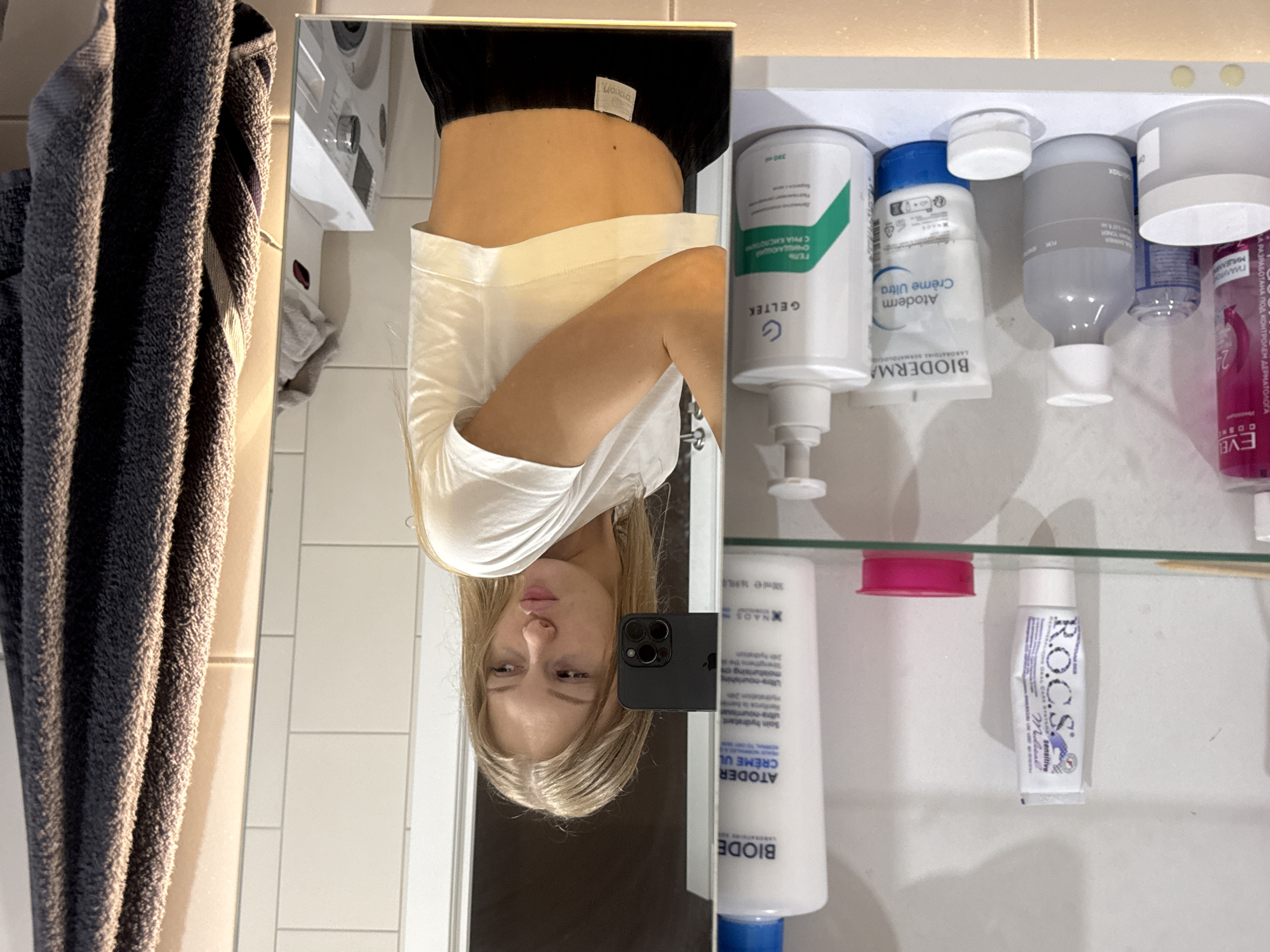 A young woman with blonde hair is taking a mirror selfie in a bathroom. She is wearing a white crop top and black pants. She is standing in front of a shelf filled with various cosmetic products, including tubes of cream and bottles of lotion. A grey towel is hanging on the right side of the frame. In the reflection of the mirror, a washing machine and tiled walls are visible. The overall scene suggests a personal grooming or self-care moment. The specific location in the city of Perm, Russia is not discernible from the image.