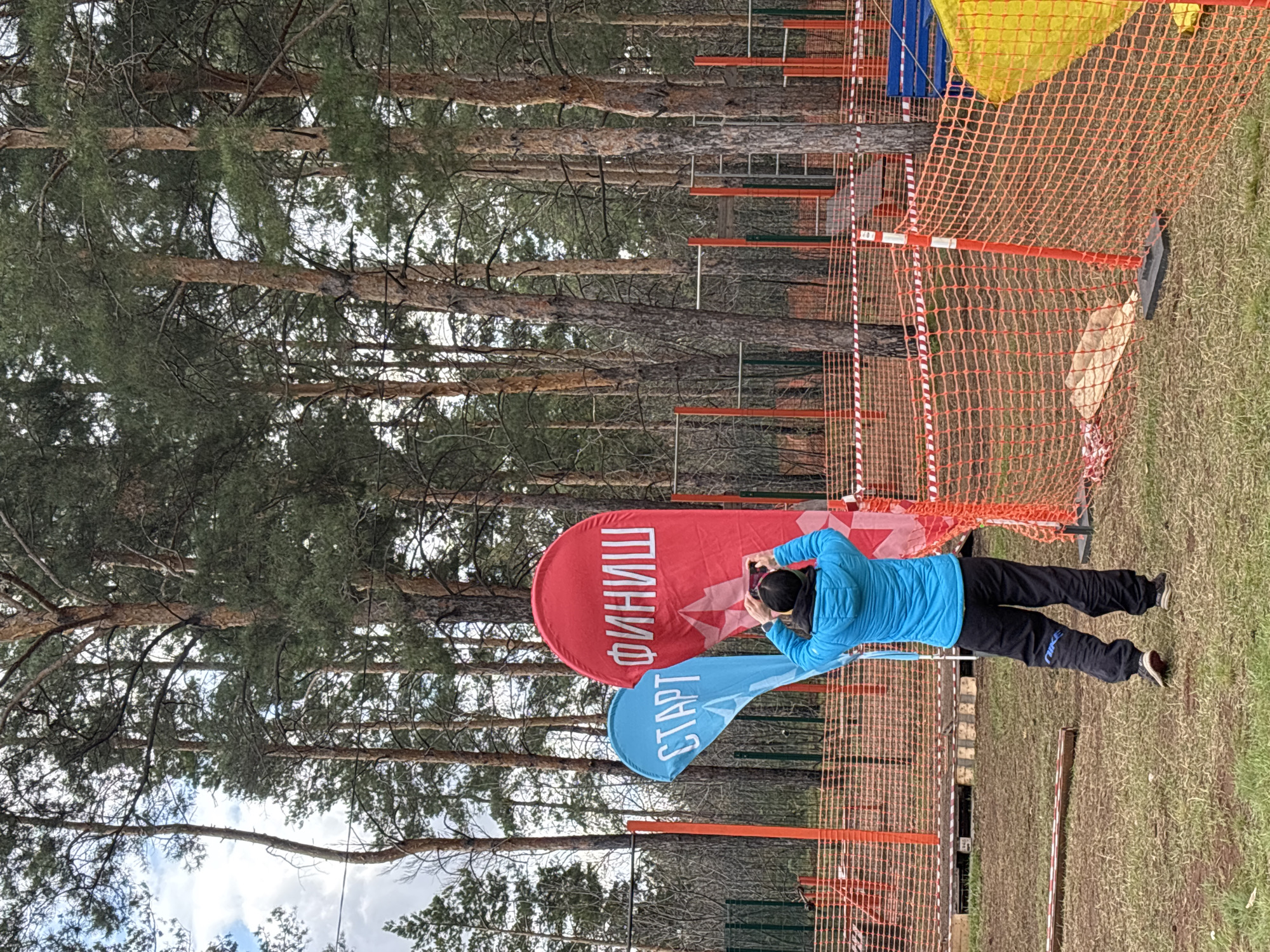 The image shows an outdoor scene near a pine forest, possibly in a park or recreational area. A person wearing a blue jacket and black pants is standing near a blue "START" flag and a red "FINISH" flag (in Russian). The person is holding a mobile phone, seemingly taking a picture of the flags or the surrounding area. The area is marked off by orange safety netting and red and white striped tape. The location is the city of Orenburg, Russia.