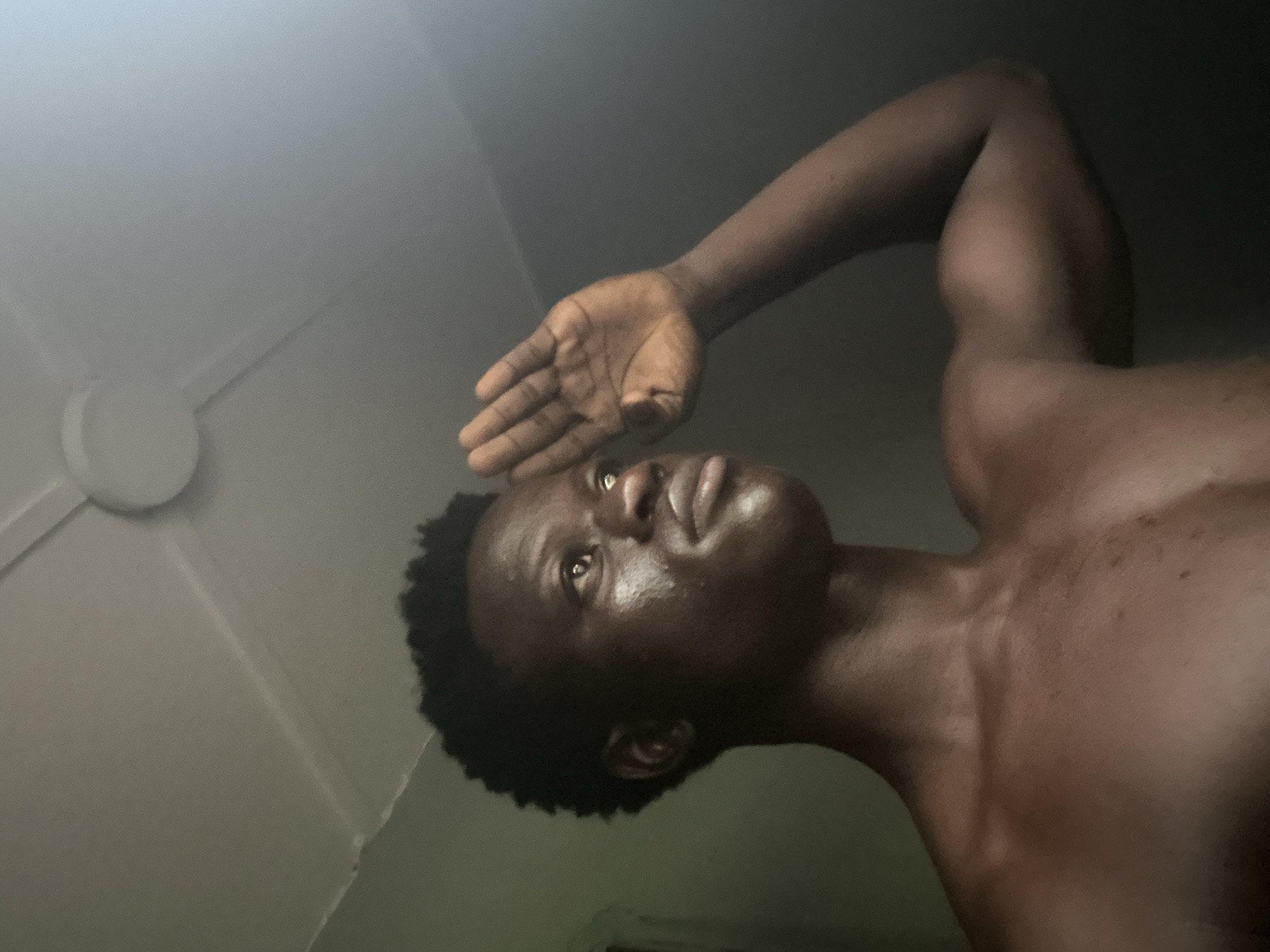 The image features a close-up, low-angle shot of a young Black male. He is shirtless and his skin appears to be moist, possibly from sweat. His right arm is raised, with his hand positioned above his forehead, as if shielding his eyes from a bright light or looking up at something. His gaze is directed upwards and slightly to the left.
The background is dimly lit and appears to be an indoor setting. A white ceiling fan is visible in the upper left portion of the image, with its blades extended. The overall lighting suggests it could be daytime, possibly with the sun's rays entering the room or a strong artificial light source. The mood conveyed is one of contemplation or perhaps searching. The subject's expression is serious.
The location context provided is Badagry, Nigeria, which, combined with the indoor setting and the presence of a ceiling fan, aligns with typical domestic environments in that region. There is no visible text in the image.