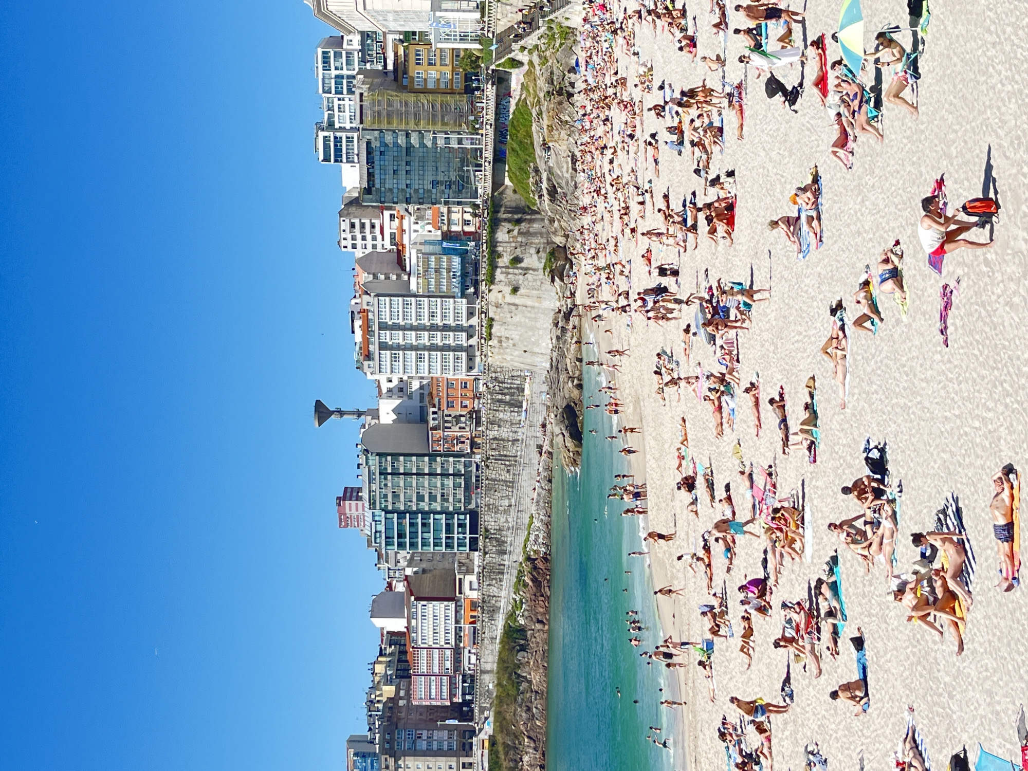 This is an aerial, high-angle photograph of a crowded beach in A Coruña, Spain. The image captures a vibrant scene on a sunny day, with a clear blue sky dominating the upper left portion of the frame. The beach is filled with numerous people relaxing on the sand, sunbathing, and enjoying the water. They are spread out across the sandy expanse, with many lying on towels or beach chairs, some under umbrellas. The ocean water is a striking turquoise color, with small waves lapping at the shore.
Along the edge of the beach, a series of tall, modern buildings rise up, featuring glass facades and balconies. These buildings suggest an urban setting directly adjacent to the beach. A rocky embankment with some greenery separates the beach from the buildings. The people are dressed in swimwear, indicating a summer day with warm weather. The presence of so many individuals enjoying the beach suggests a popular tourist destination or a well-frequented local spot during peak season. The sunlight casts distinct shadows on the sand, indicating it is likely midday or early afternoon. There is no visible text in the image.