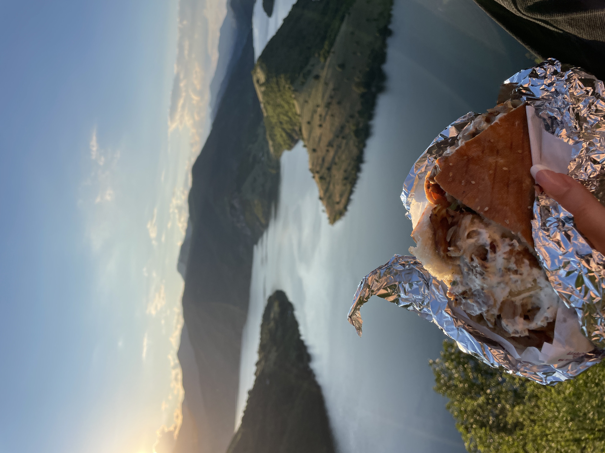 A person's hand is holding a large sandwich wrapped in foil, in the foreground. The sandwich is partially unwrapped, revealing a filling of meat, sauces, and vegetables. In the background, a panoramic view of the Arda River (also known as the Arda Reservoir) is visible, with rolling, tree-covered hills surrounding it. The sun is low in the sky, casting a warm, golden light and creating a sense of a sunset or sunrise. The sky above is a clear blue with a few wispy clouds. The location is identified as Borovitsa, Bulgaria.