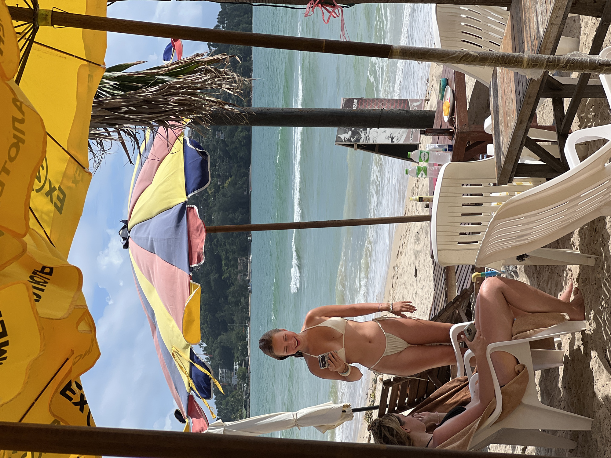 An eye-level, outdoor, medium shot captures a young woman standing on a sandy beach, holding a phone and smiling. She wears a light-colored bikini. To her left, another woman sits in a white plastic chair, facing left, also looking at her phone. In the background, the ocean stretches out with moderate waves under a cloudy sky. Beach umbrellas, chairs, and a wooden table are visible in the foreground and midground. A sign advertising massages stands near the table. The scene appears to be at a tropical beach resort.